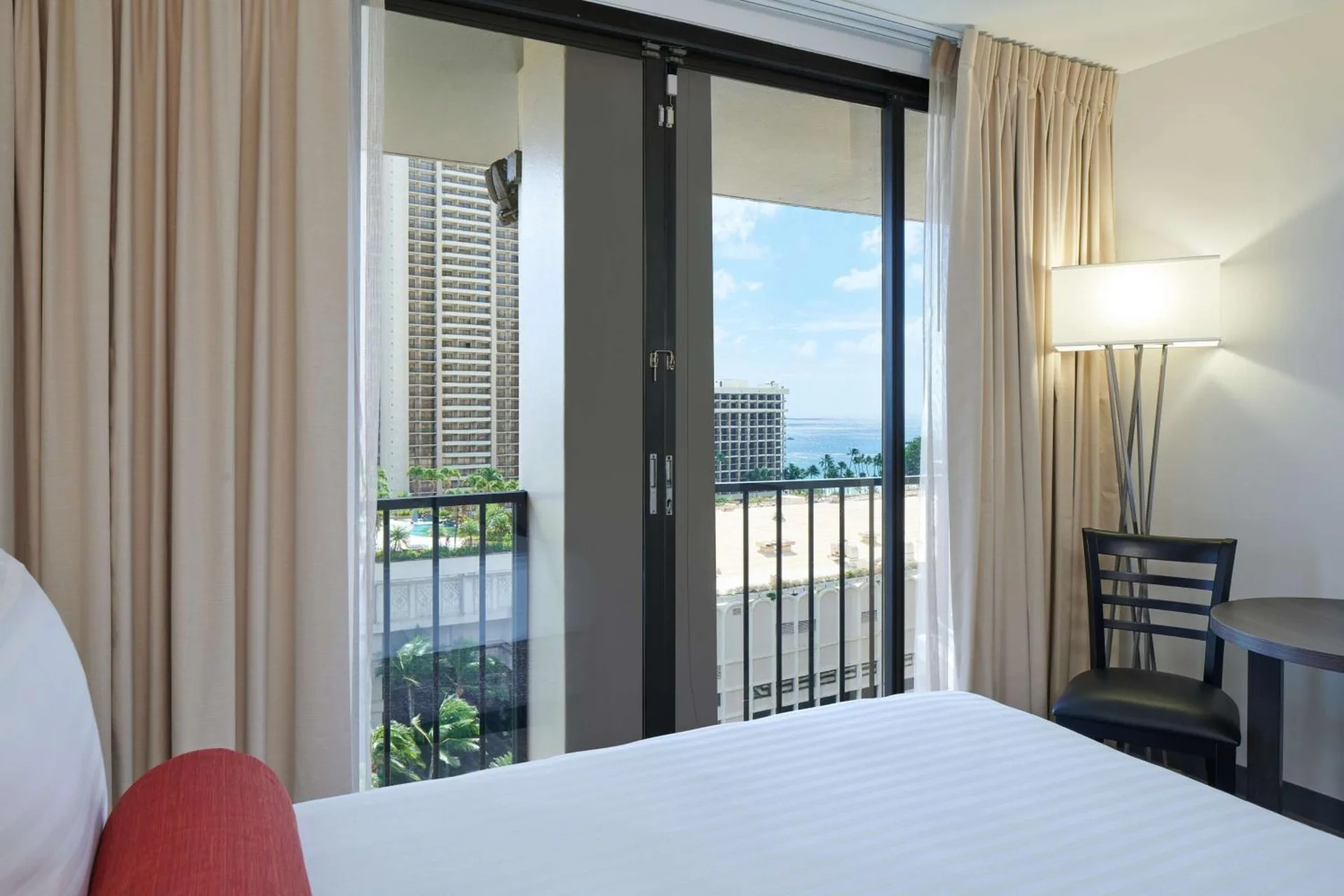 Guest room bed beside balcony doors with ocean glimpse at Aqua Palms Waikiki.