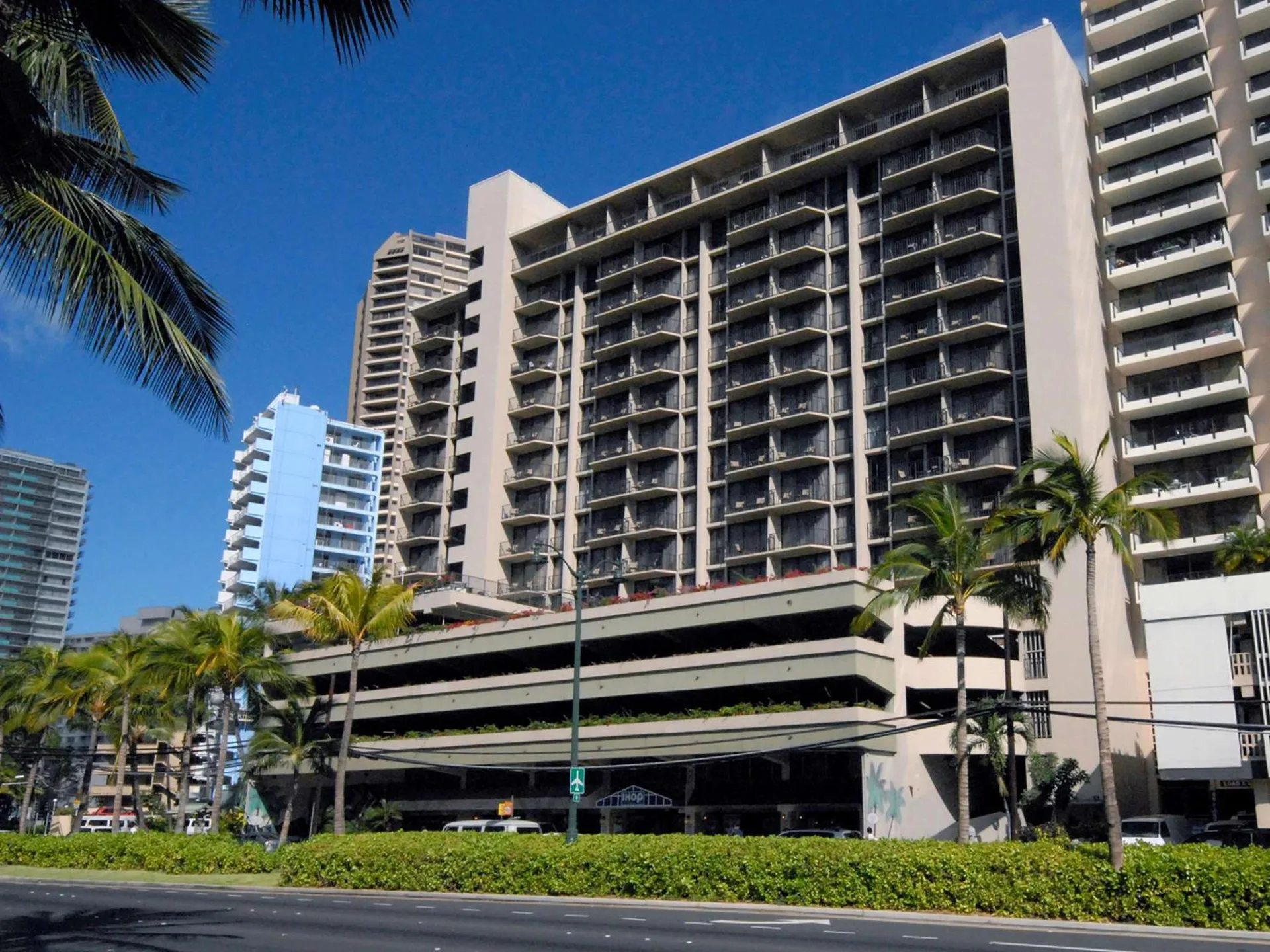 Hotel tower with stacked balconies and palm-lined street at Aqua Palms Waikiki.