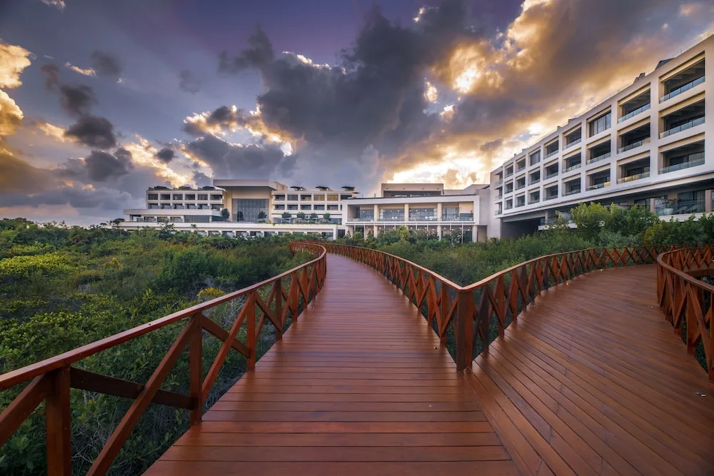Wooden walkway through greenery toward resort buildings at Atelier Playa Mujeres.
