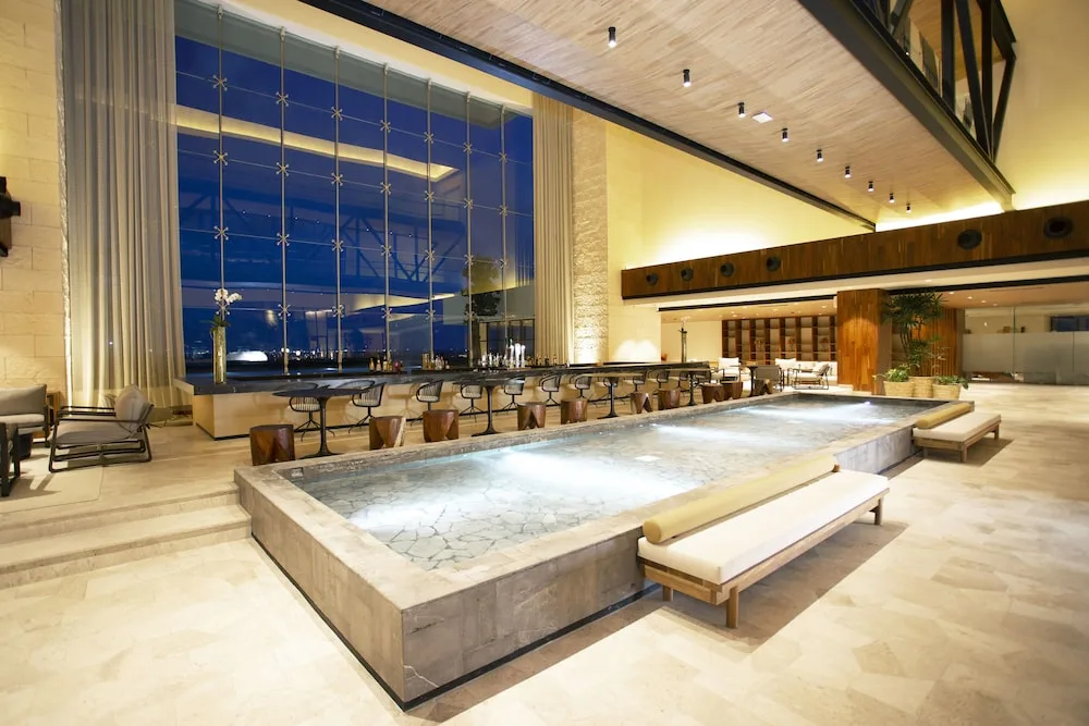 Night lobby bar at Atelier Playa Mujeres with illuminated reflecting pool and tall windows.