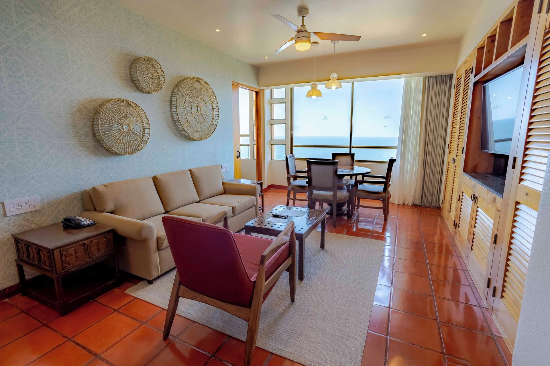 Suite living room with sofa, armchair, dining table, TV cabinet, and ocean windows at Club Regina Puerto Vallarta.