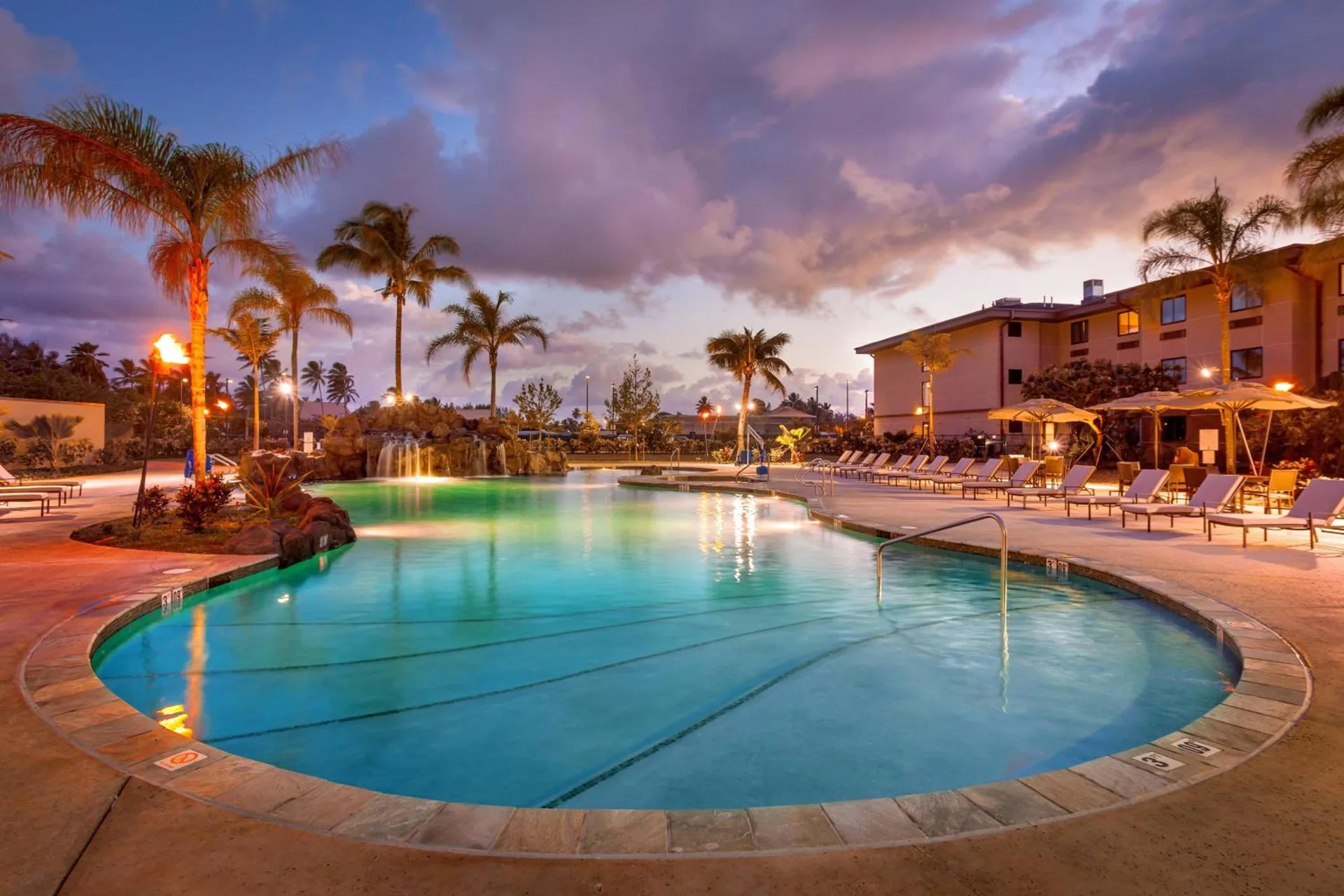 Outdoor pool at Courtyard Oahu North Shore at dusk with lounge chairs, palms and resort buildings.