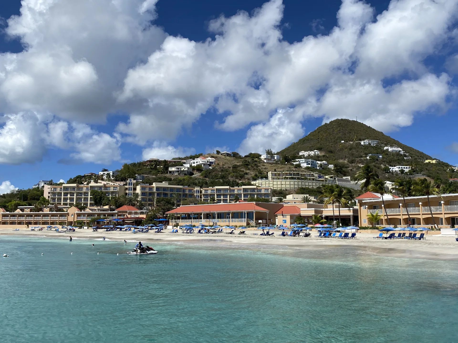 Divi Little Bay Beach Resort lines a turquoise beach below green hills and bright clouds.