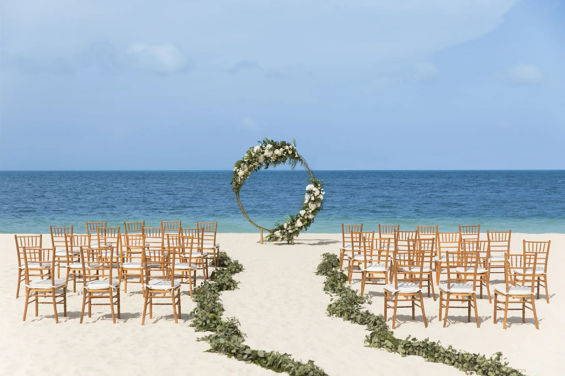 A beach wedding aisle with wooden chairs and a floral circle arch at Excellence Playa Mujeres.