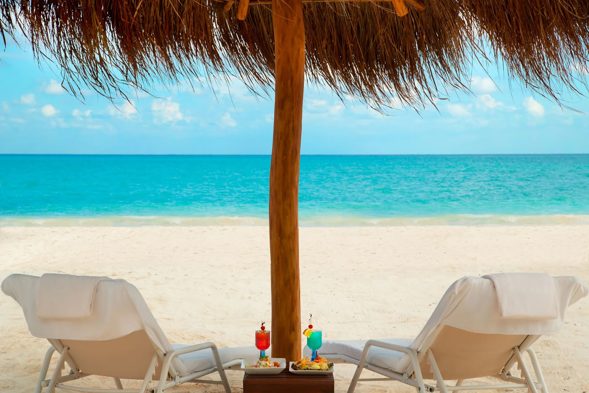 Two beach loungers with drinks sit under a palapa facing the sea at Fairmont Mayakoba.