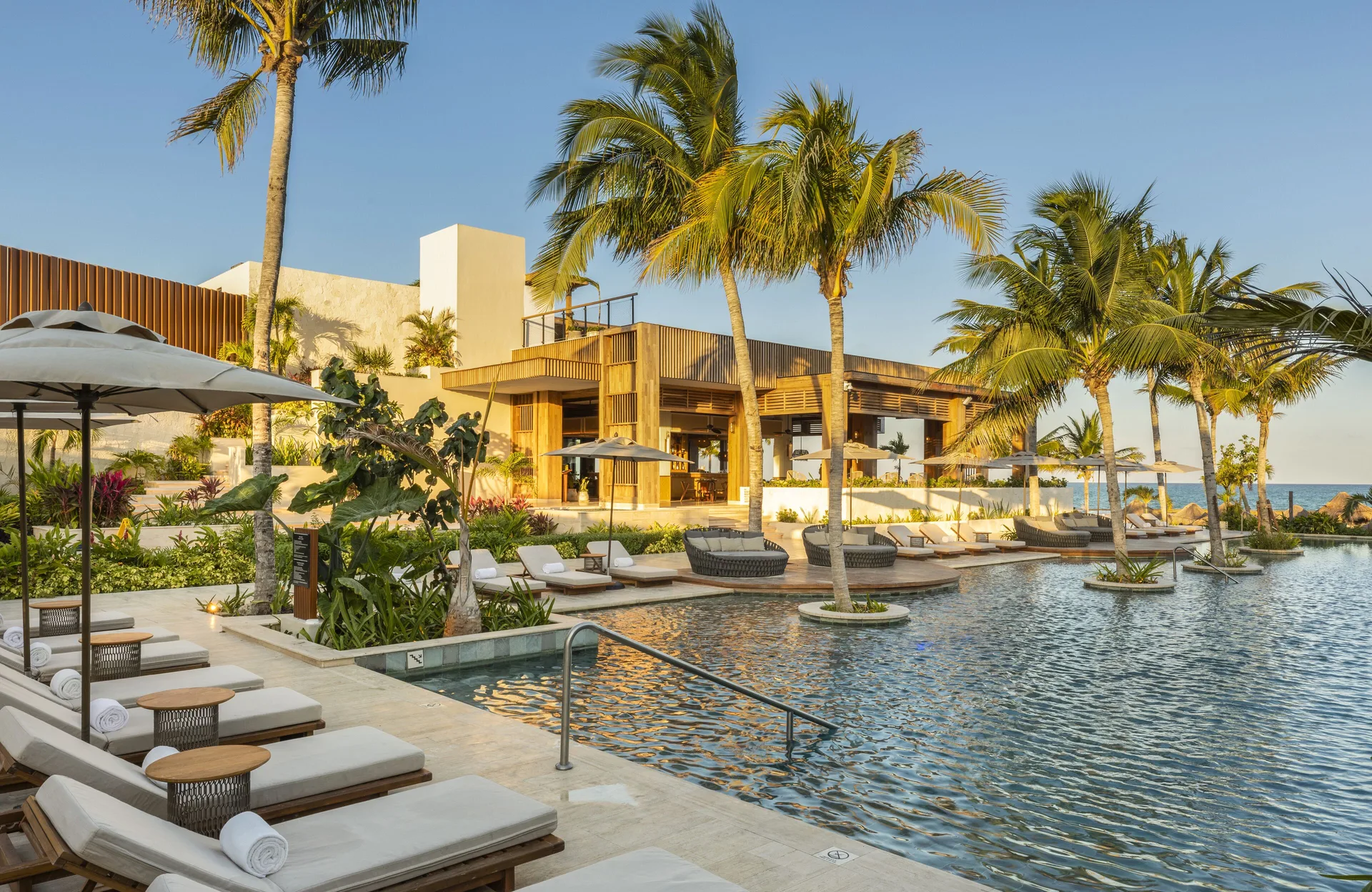 Resort pool with loungers, palm trees, umbrellas, and ocean beyond at Fairmont Mayakoba.