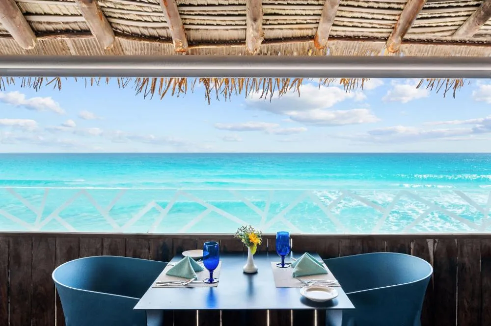 Oceanfront table under a palapa roof with blue glasses at GR Solaris Caribe.