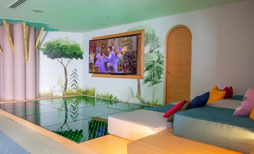 Kids' lounge at Grand Velas Los Cabos with a wall TV, mural, cushions, and lighted floor.