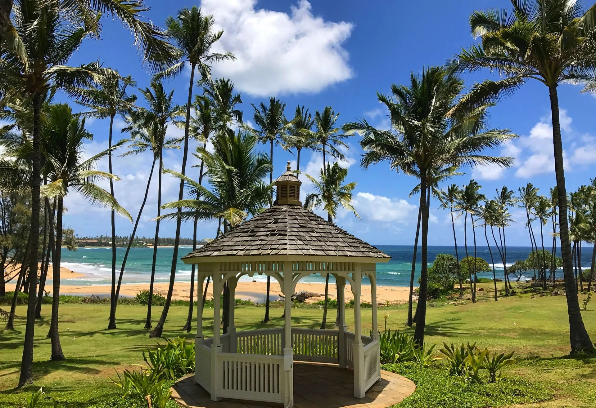 White gazebo, palms, lawn, and beach at Hilton Garden Inn Kauai Wailua Bay.