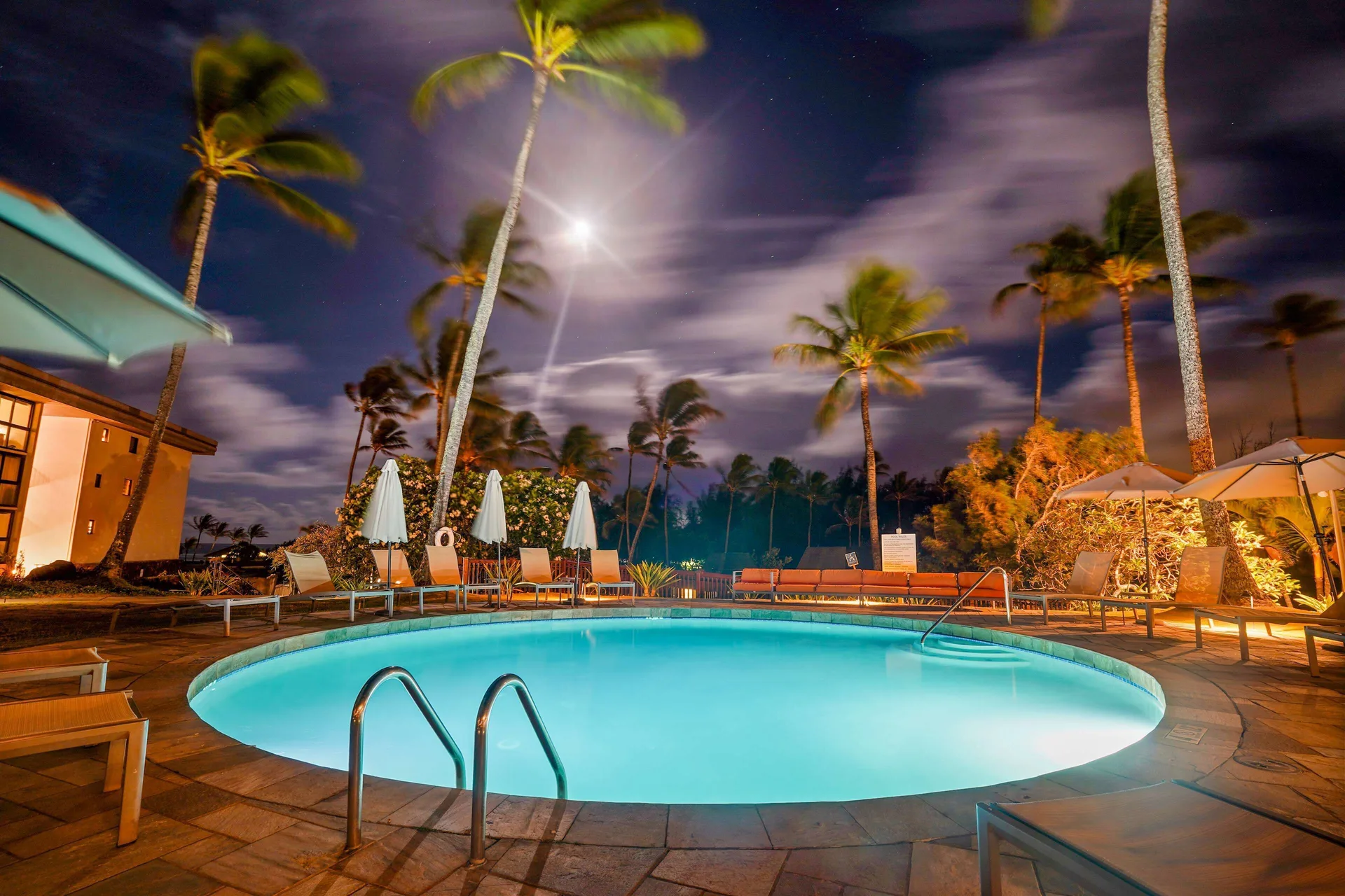 Round illuminated pool with lounge seating and palms at Hilton Garden Inn Kauai Wailua Bay.