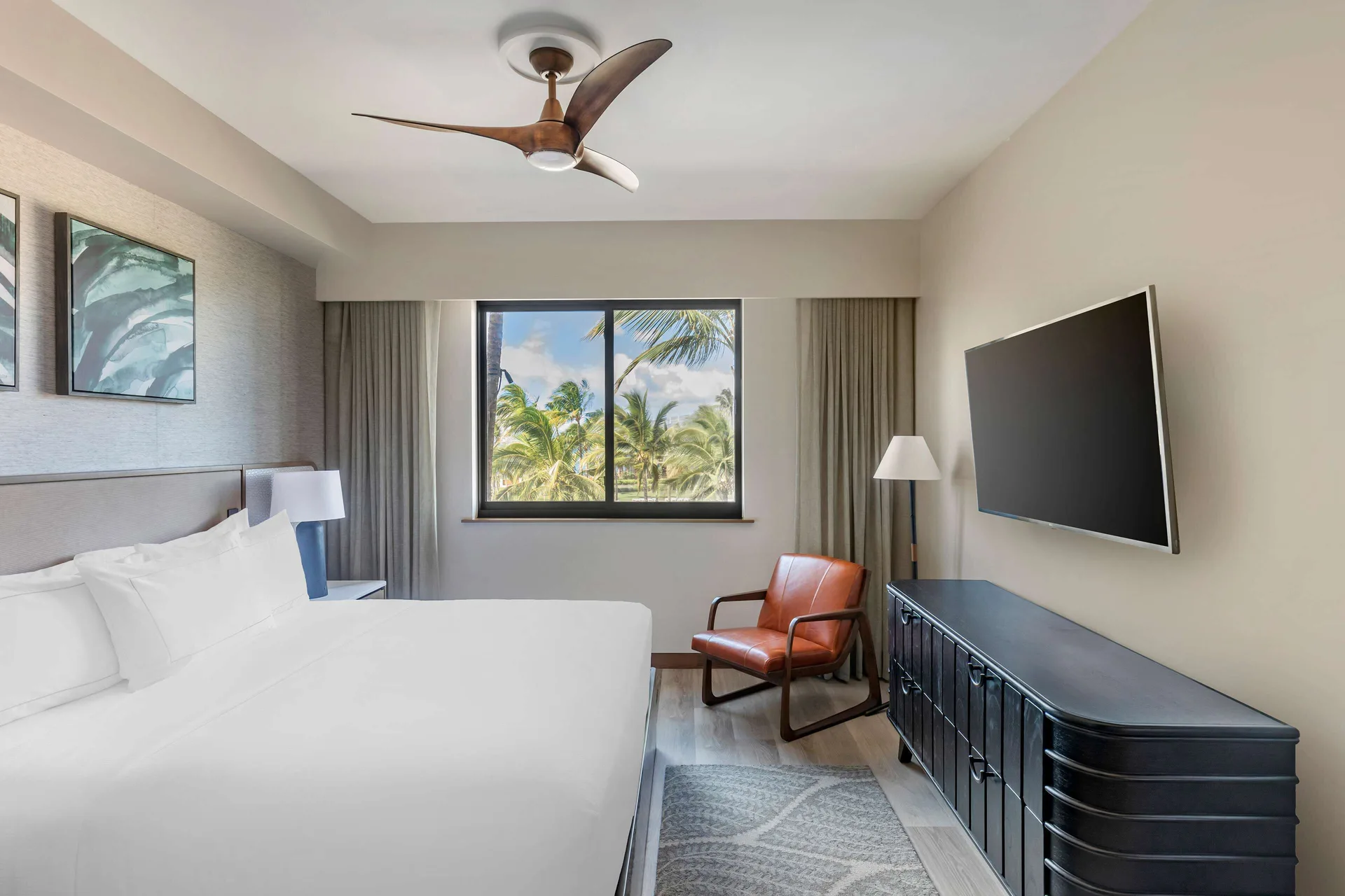 King bedroom with wall TV, leather chair, and palm-framed window at Hilton Grand Vacations Club Maui Bay Villas.