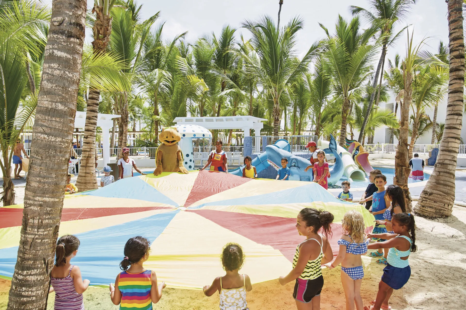 Children play with a colorful parachute in the palm-shaded kids area at Hotel Riu Bambu.