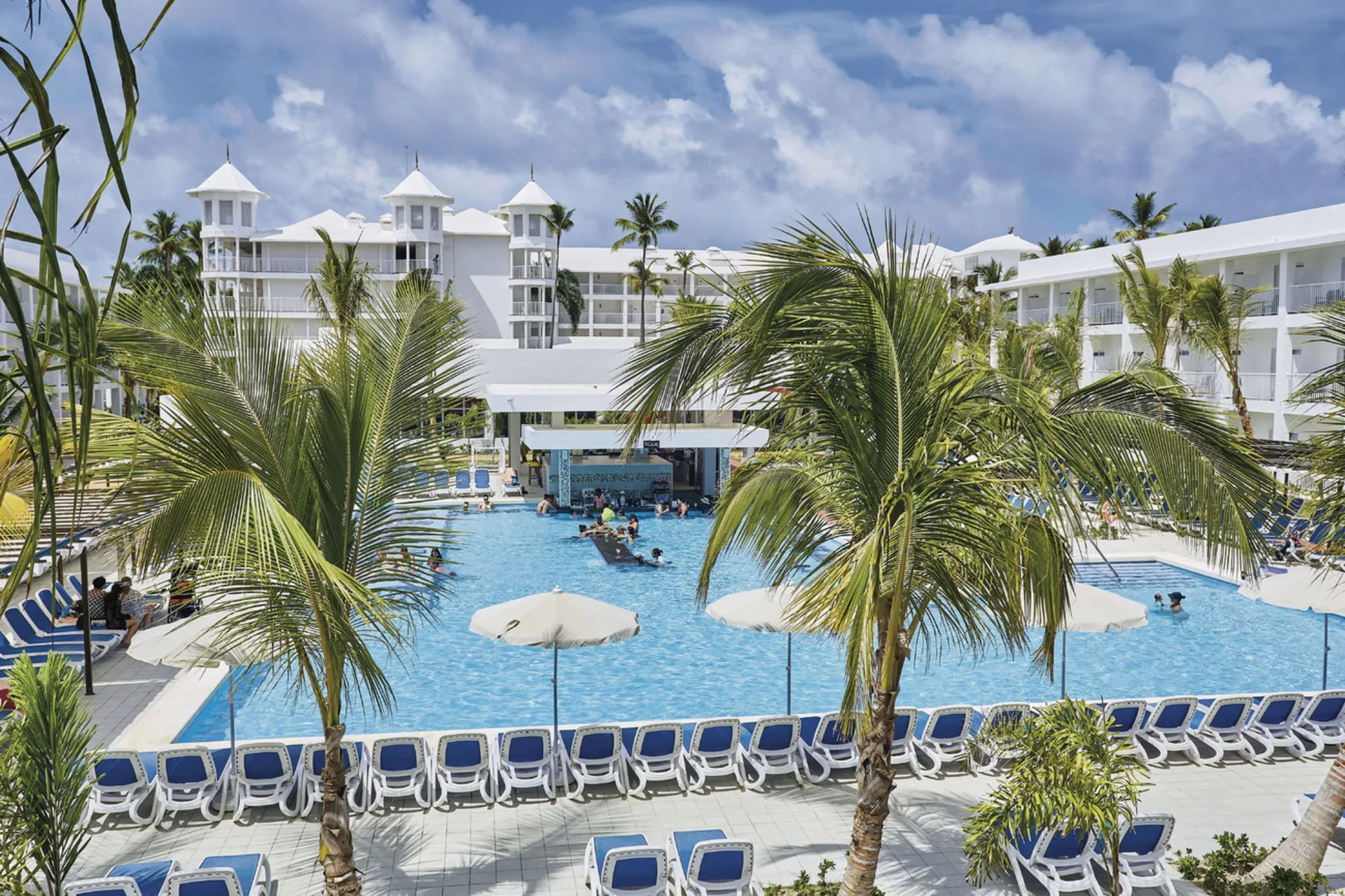 Palm-framed pool with loungers, umbrellas, and white buildings at Hotel Riu Bambu.