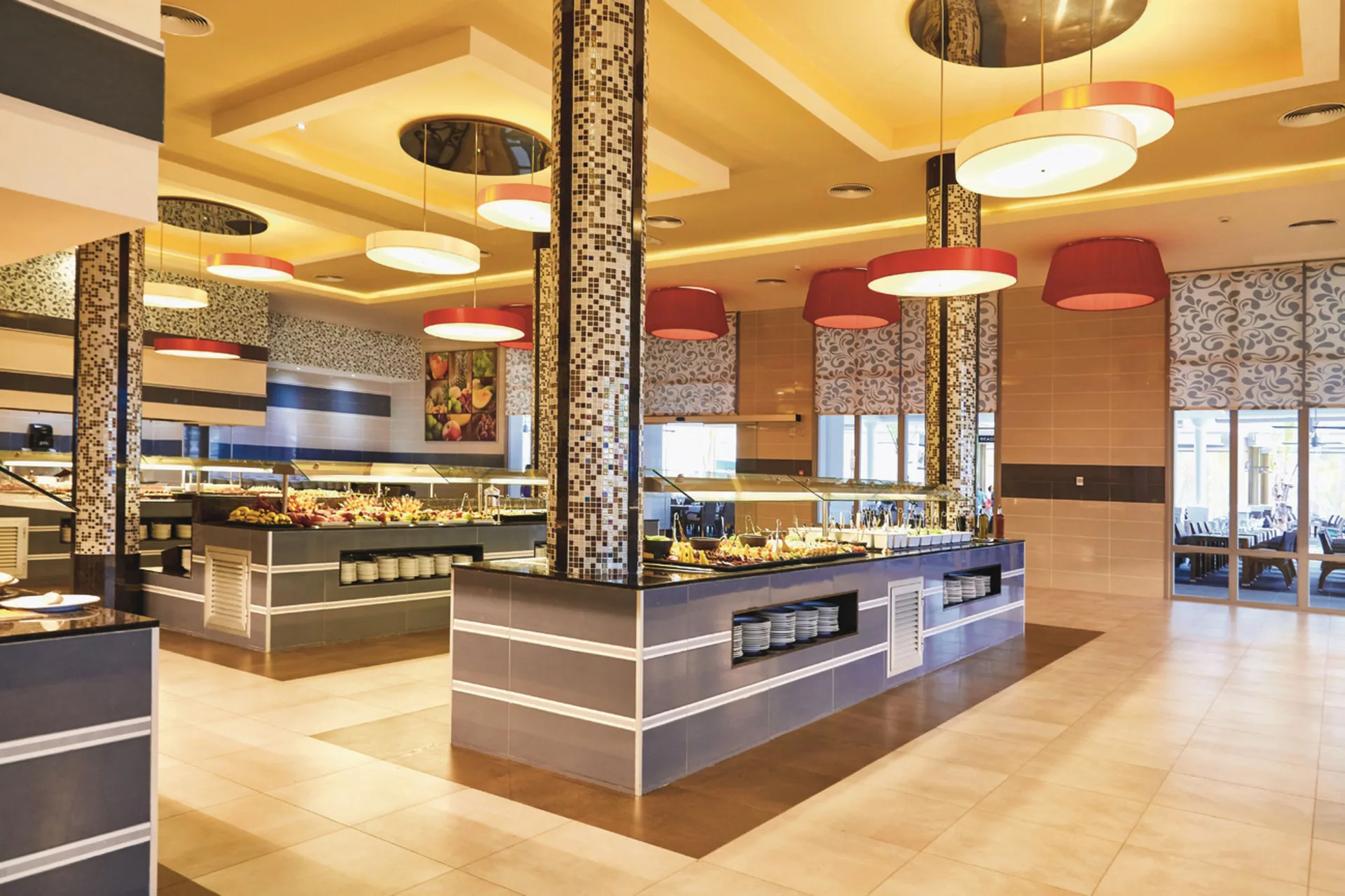 A bright buffet dining room with serving islands and pendant lights at Hotel Riu Bambu.