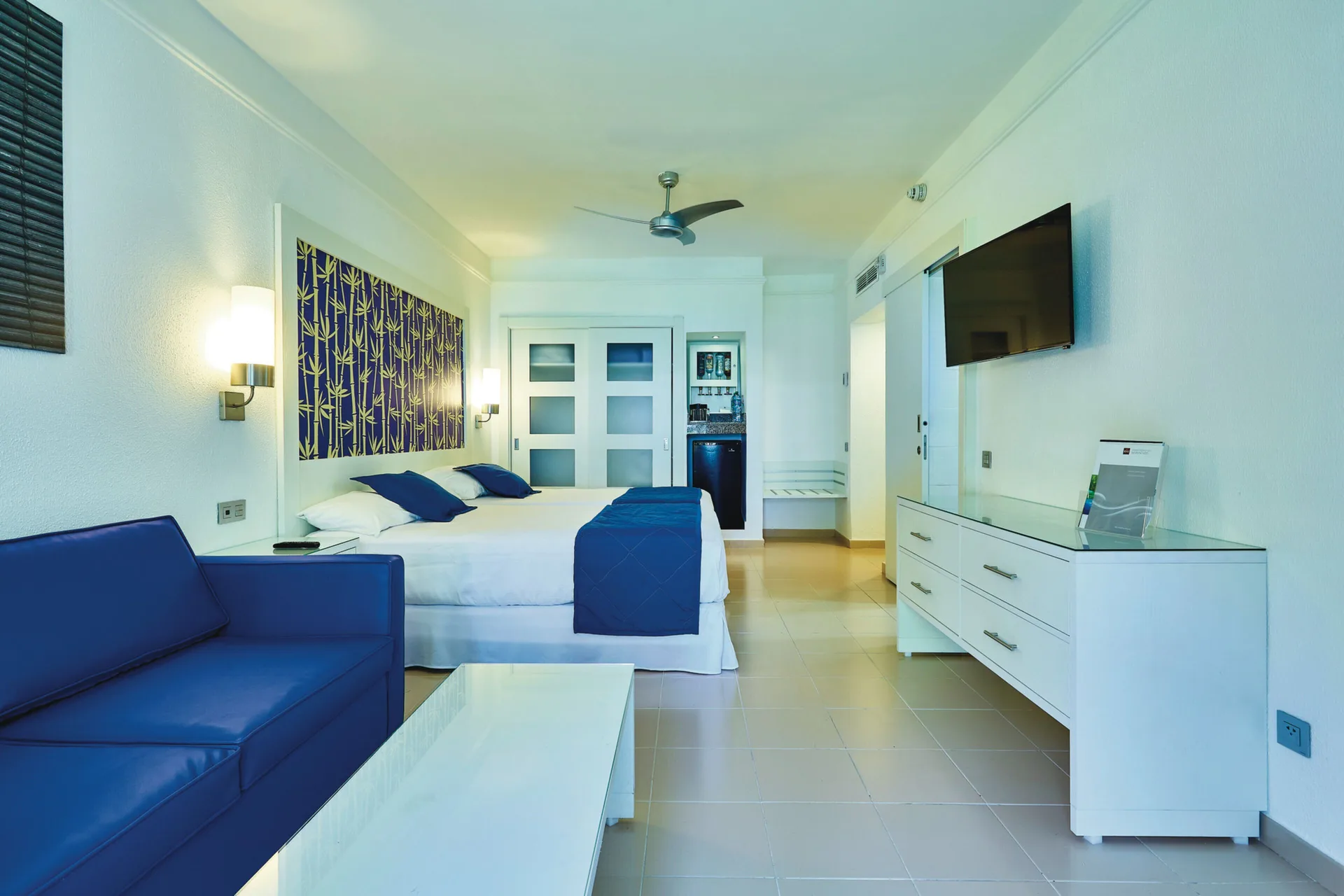 Family Room with bed, blue sofa, TV dresser, and refreshment area at Hotel Riu Bambu.