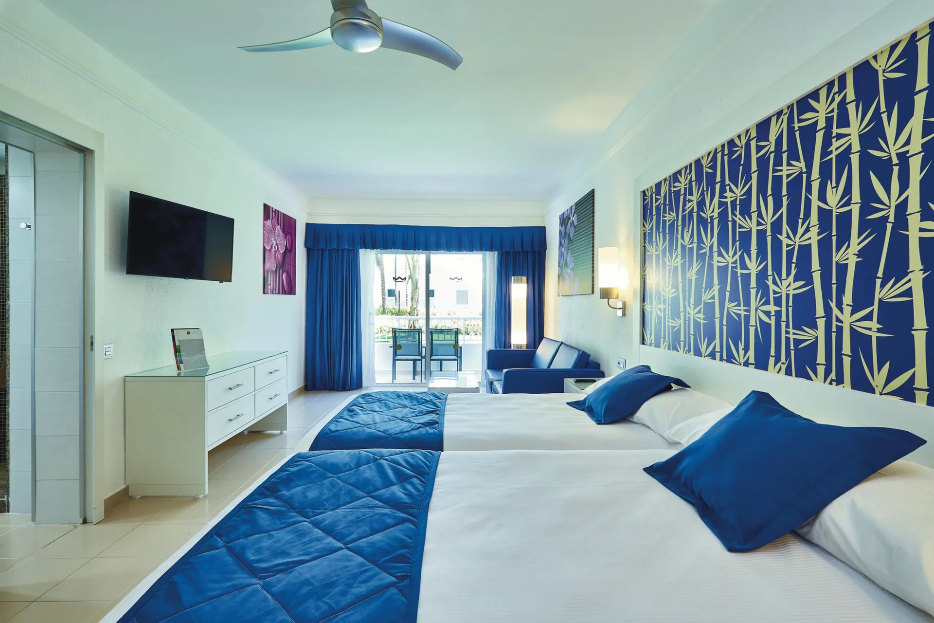 Family Room with two beds, blue sofa, TV, and balcony at Hotel Riu Bambu.