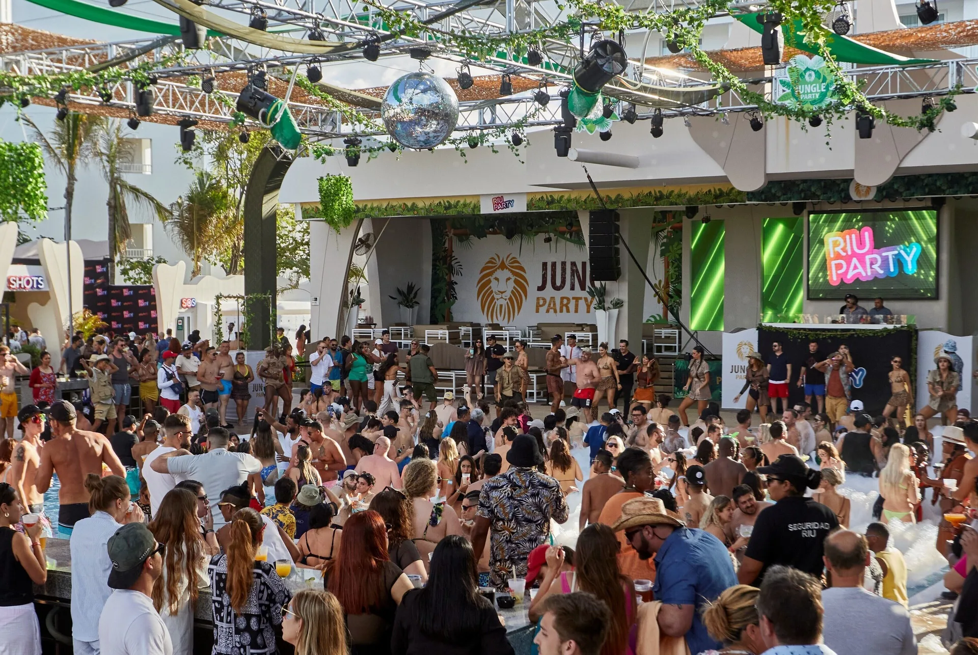 A crowded daytime pool party with stage lighting and tropical decor at Hotel Riu Cancun.