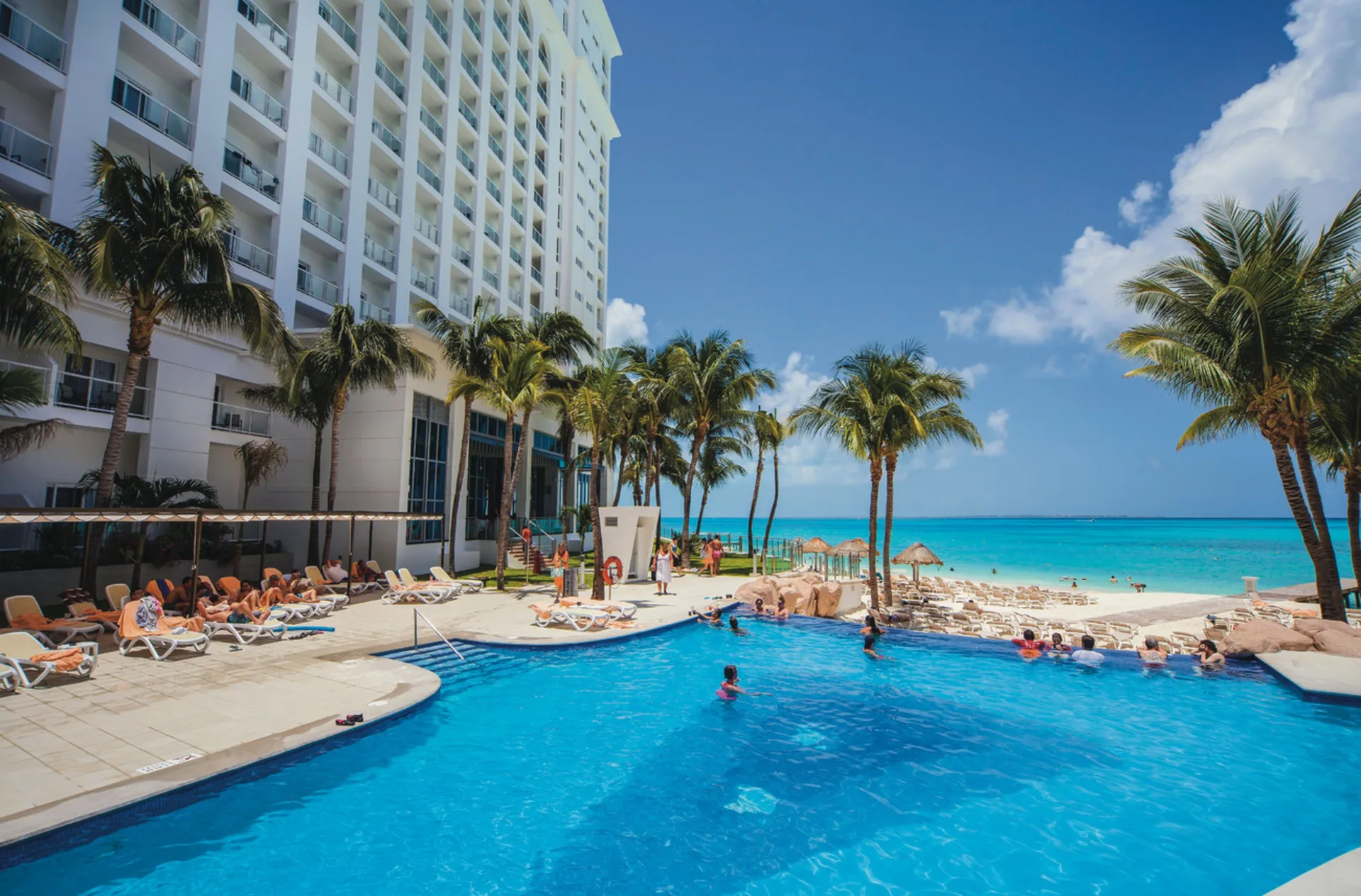 Pool area at Hotel Riu Cancun with palms, loungers, guests, and the beach beyond.