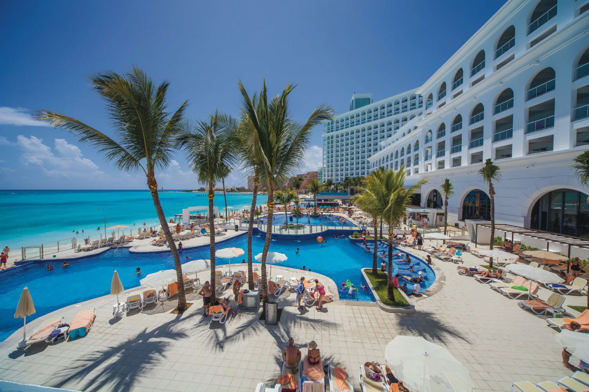 Elevated view of Hotel Riu Cancun pools, palms, loungers, and beach shoreline.