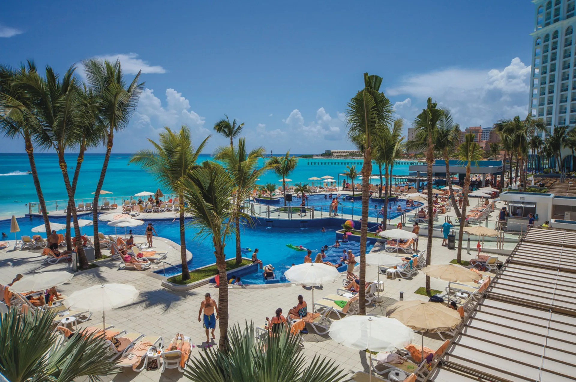 Busy pool deck at Hotel Riu Cancun with palm trees, umbrellas, loungers, and ocean views.