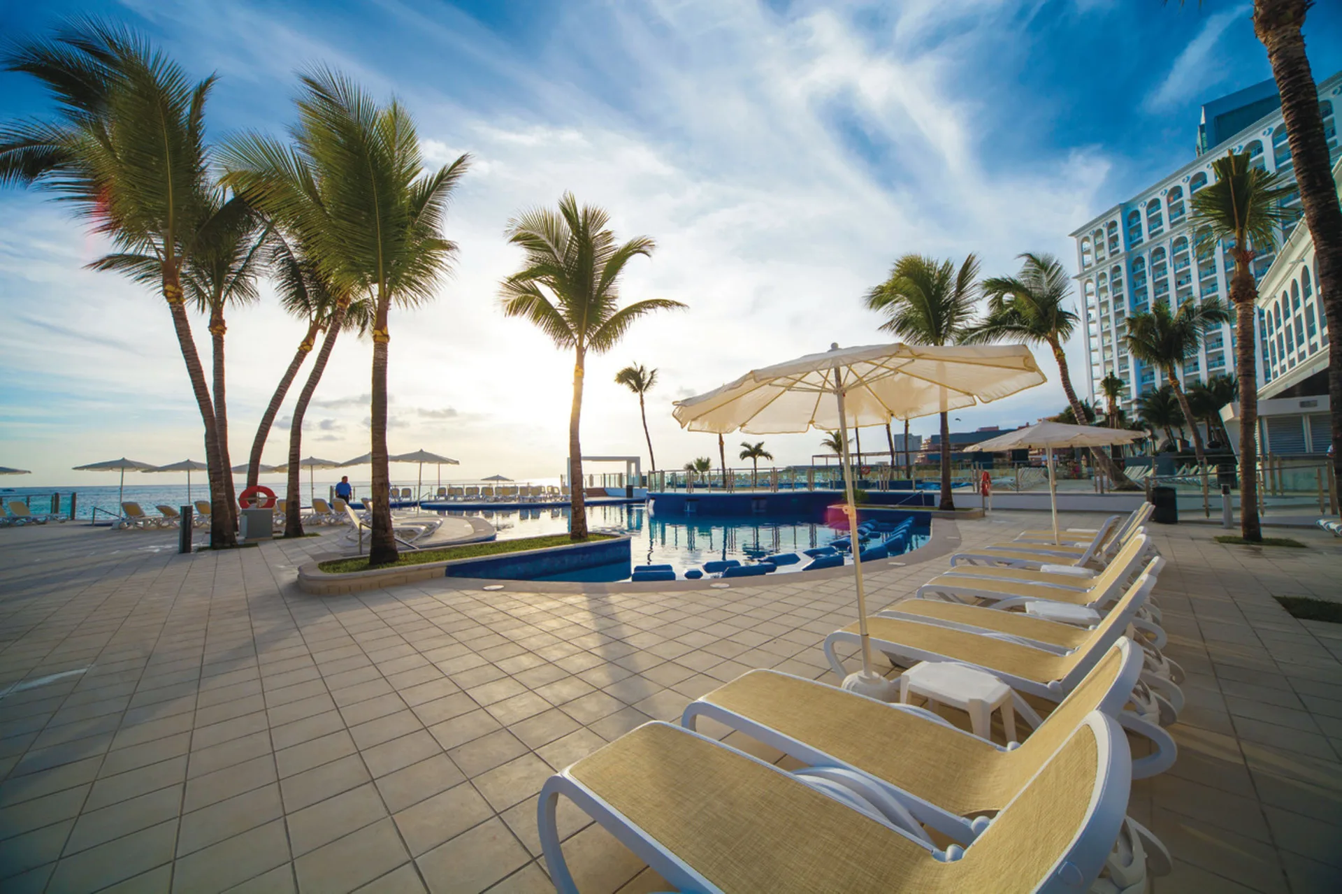 Sunlit pool deck at Hotel Riu Cancun with loungers, umbrellas, palms, and sea views.