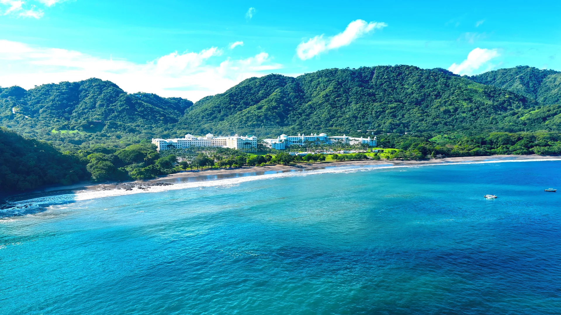 Aerial view of Hotel Riu Guanacaste on a turquoise bay below green hills.
