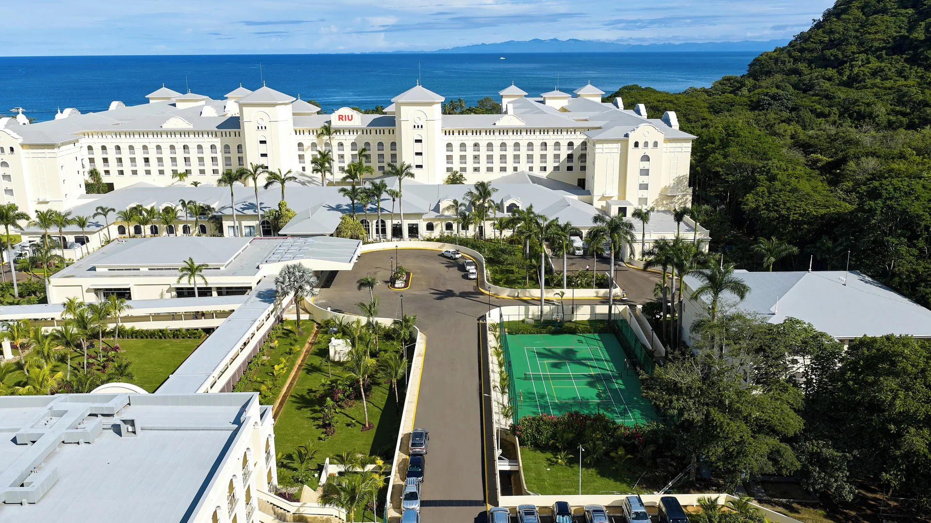 Aerial entrance view of Hotel Riu Guanacaste with driveway, tennis court, palms, and ocean beyond.