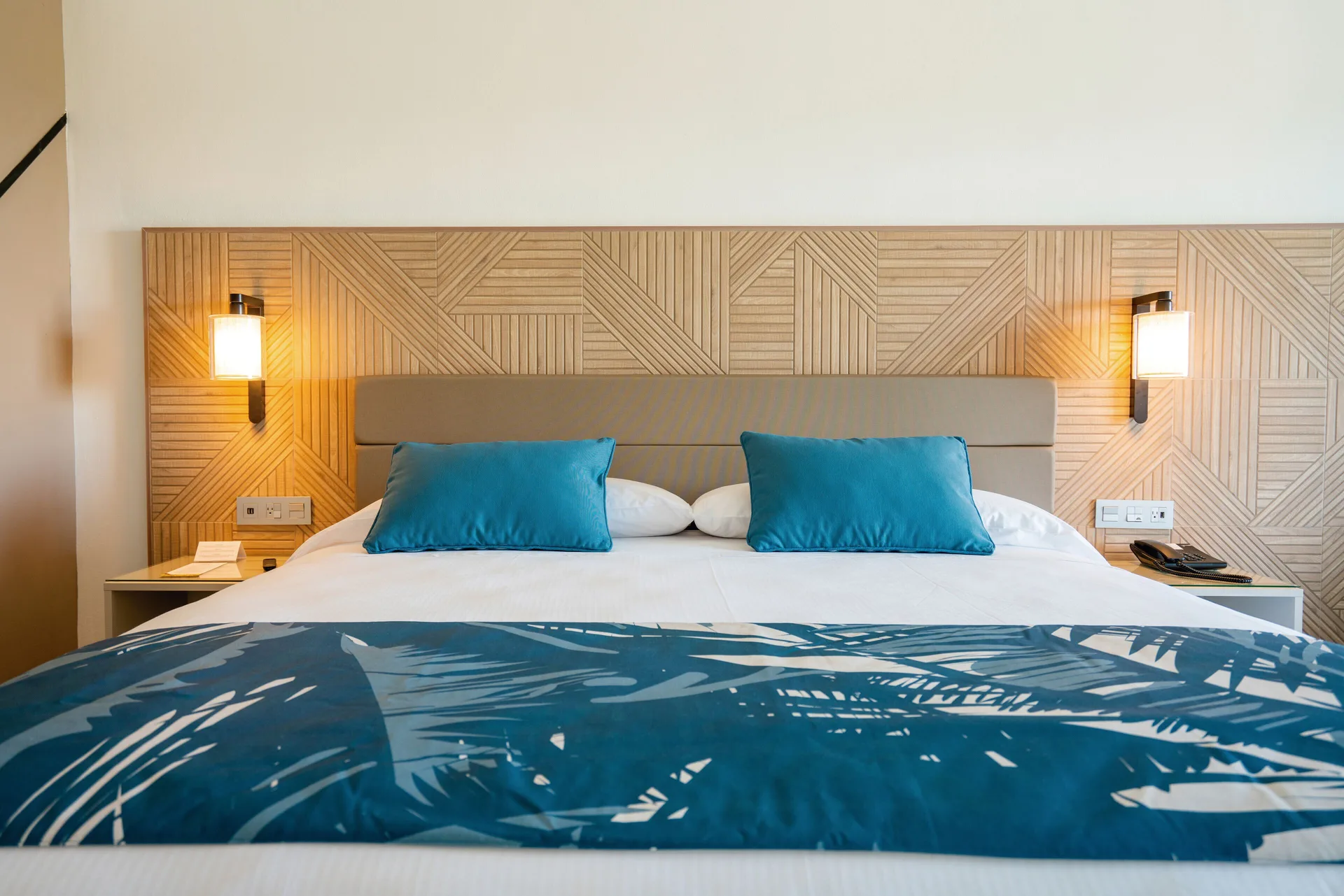 Partial Ocean View room bed with blue accents and wall lamps at Hotel Riu Latino.