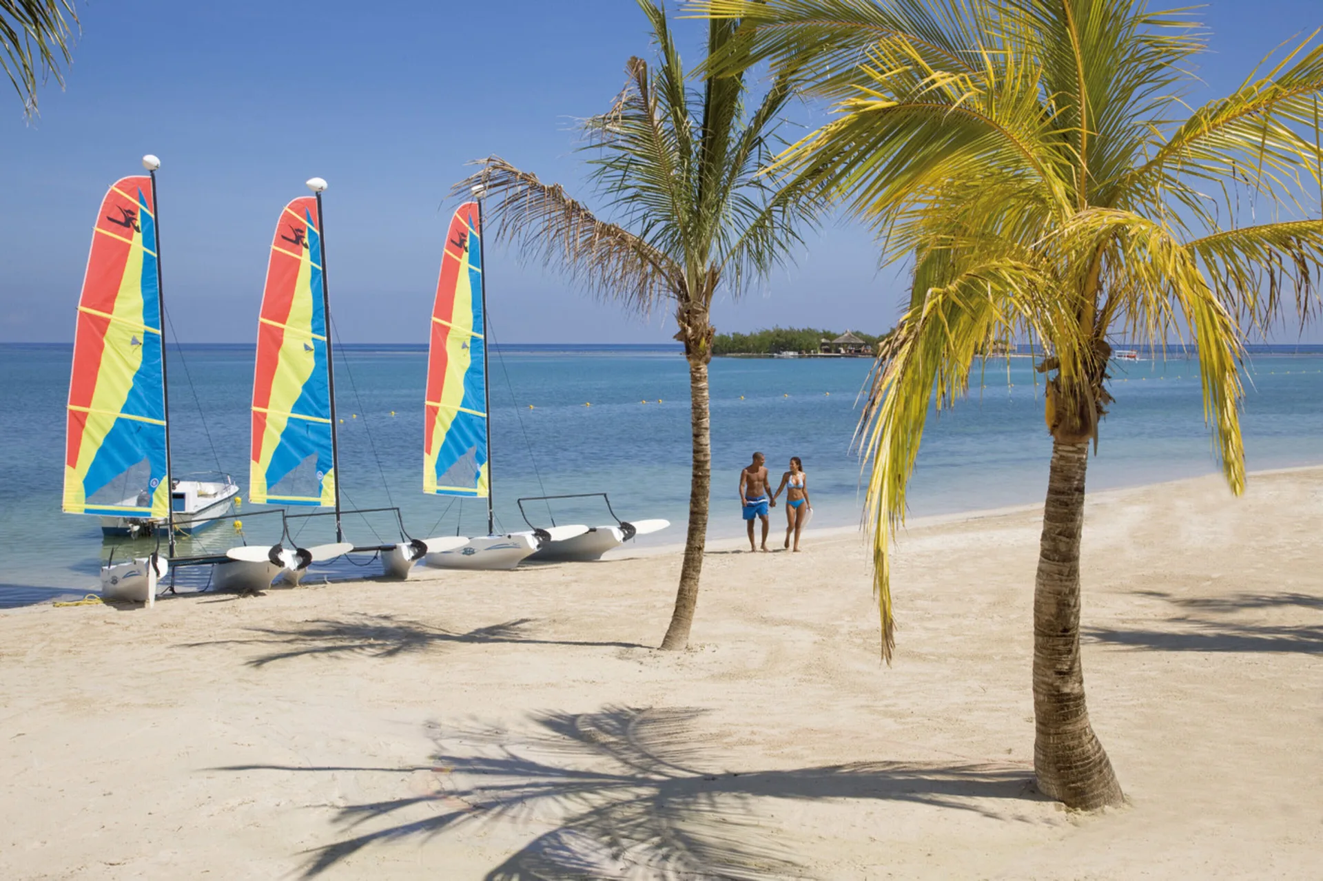 Sandy beach at Hotel Riu Montego Bay with palm trees, calm water, and small sailboats on shore.