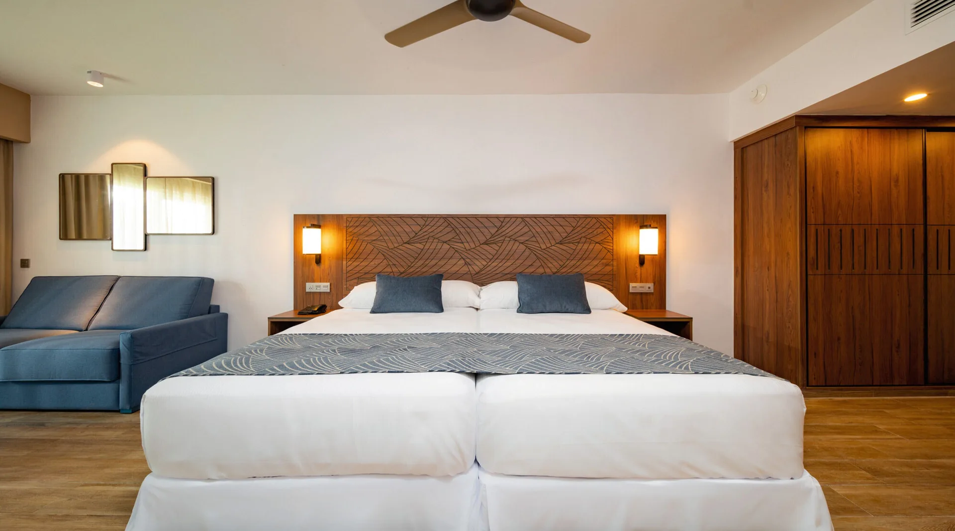 Guest room with joined beds, blue sofa, ceiling fan and wood closet at Hotel Riu Montego Bay.
