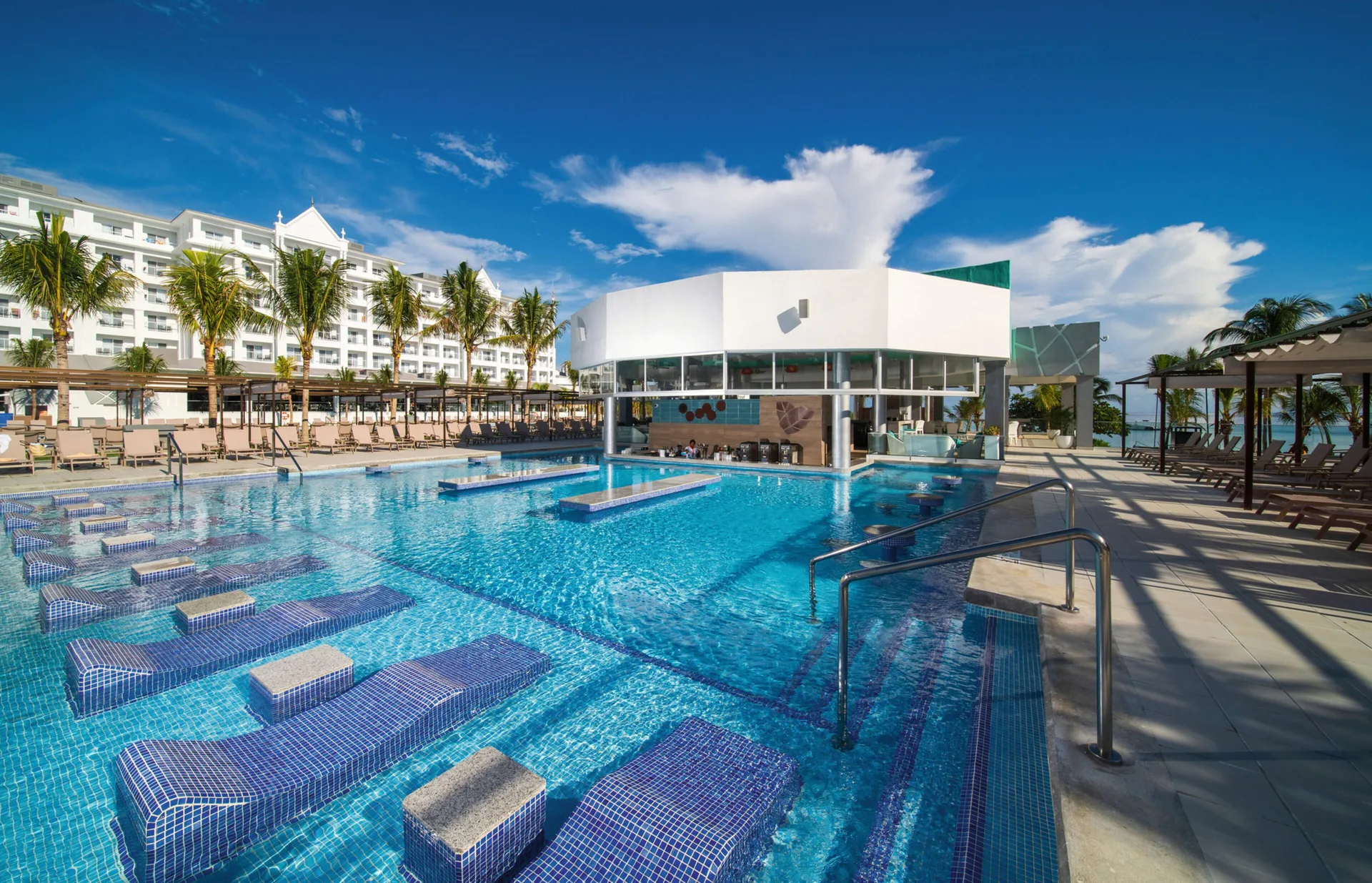 Main pool with submerged loungers, bar building and sea beyond at Hotel Riu Ocho Rios.