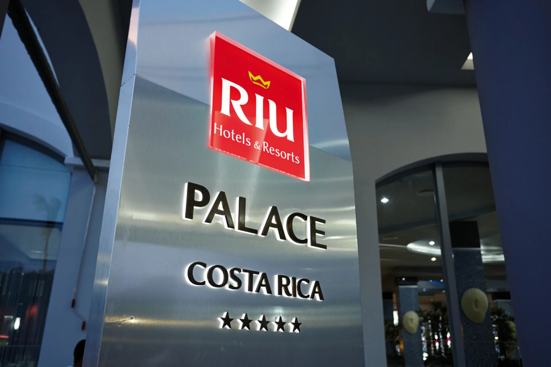 Illuminated entrance sign for Hotel Riu Palace Costa Rica beside the lobby area.