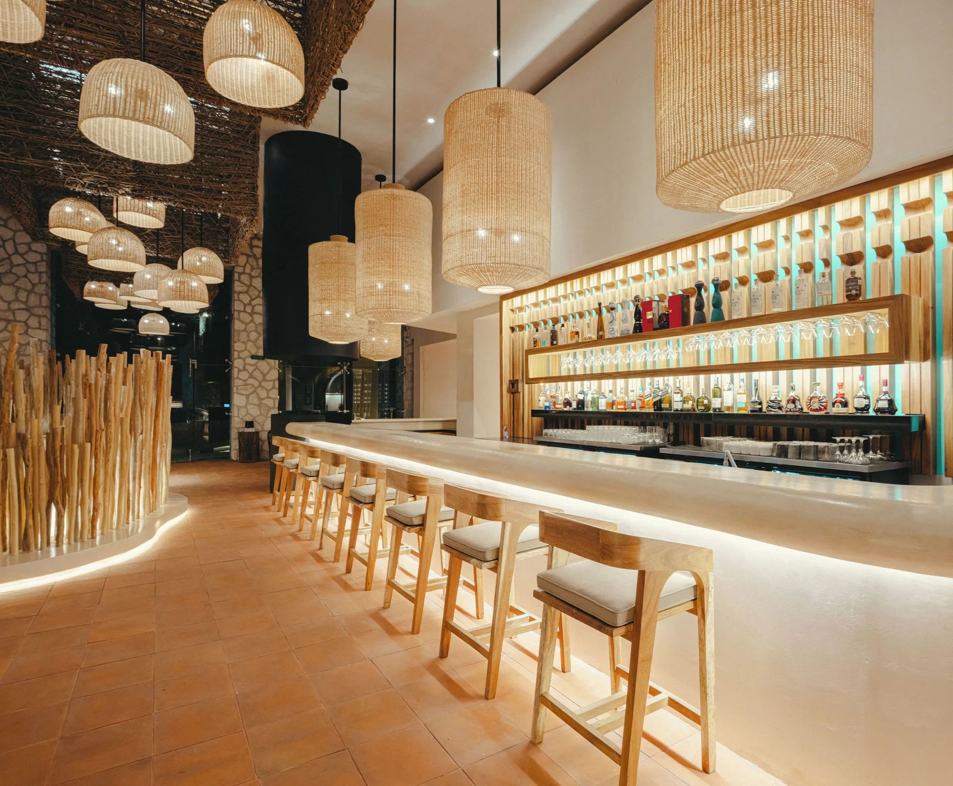 Long bar with woven pendant lights, wood stools and bottle shelves at Hotel Xcaret Arte.