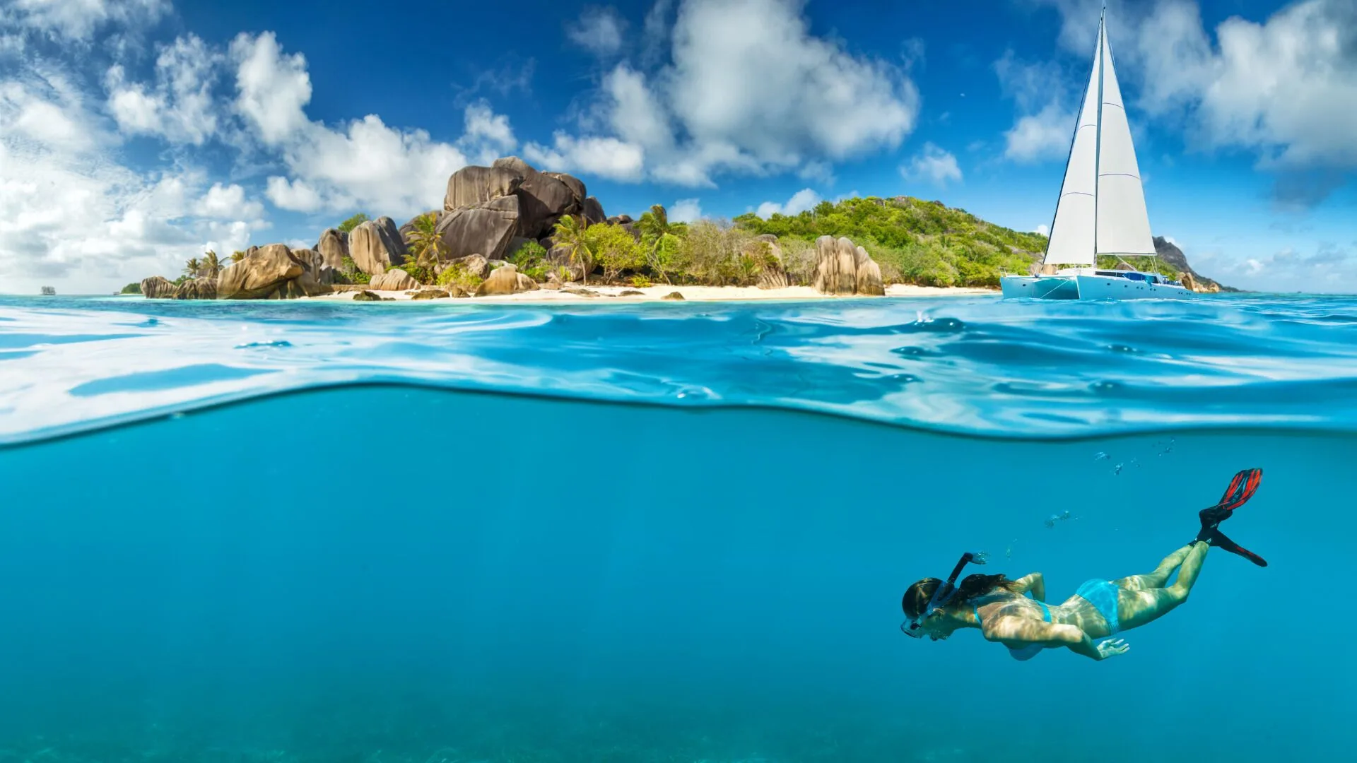 A snorkeler swims below clear blue water near a sailboat and rocky island at Jolly Beach Antigua.