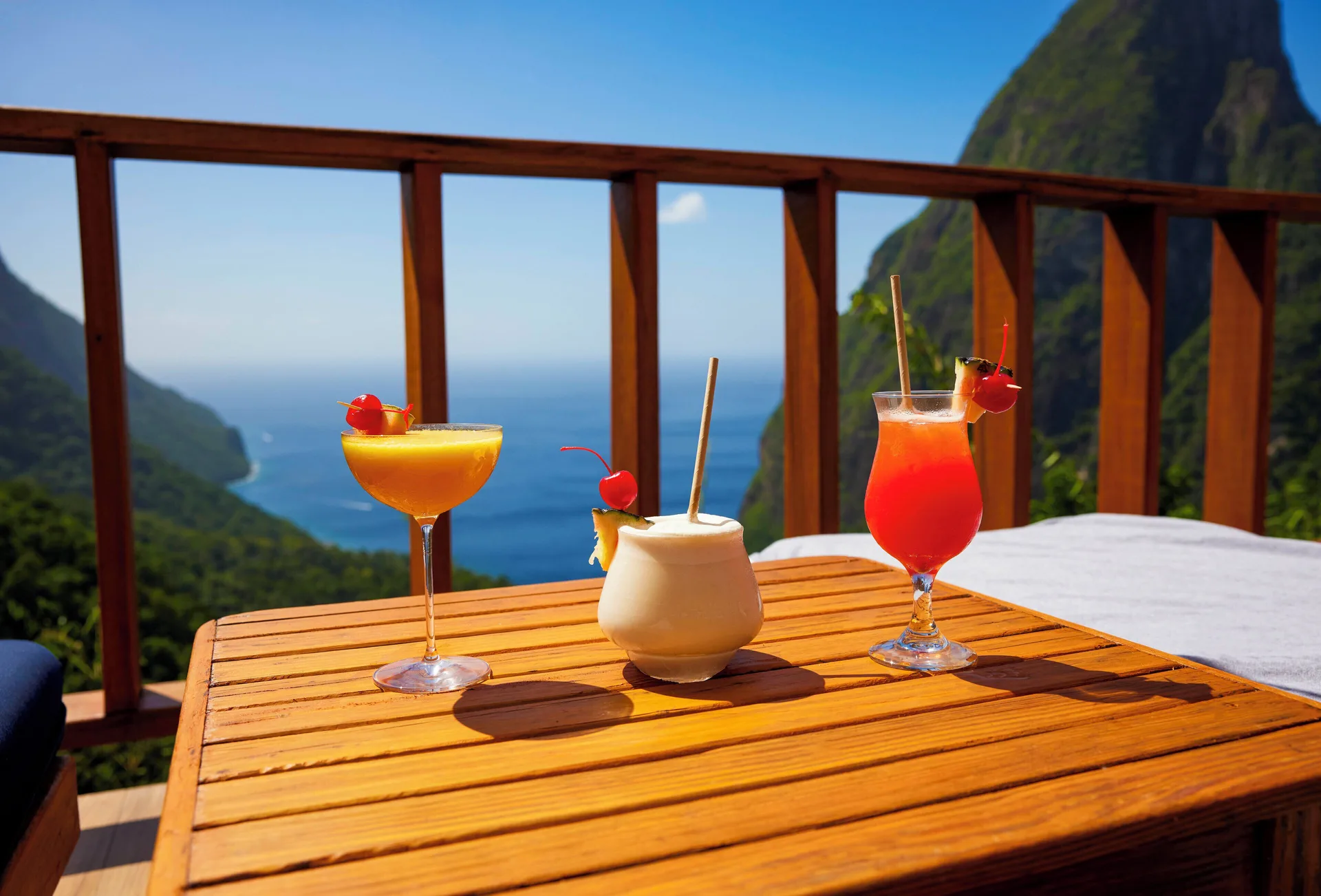 Three tropical cocktails sit on a wooden table with Piton and sea views at Ladera Resort.