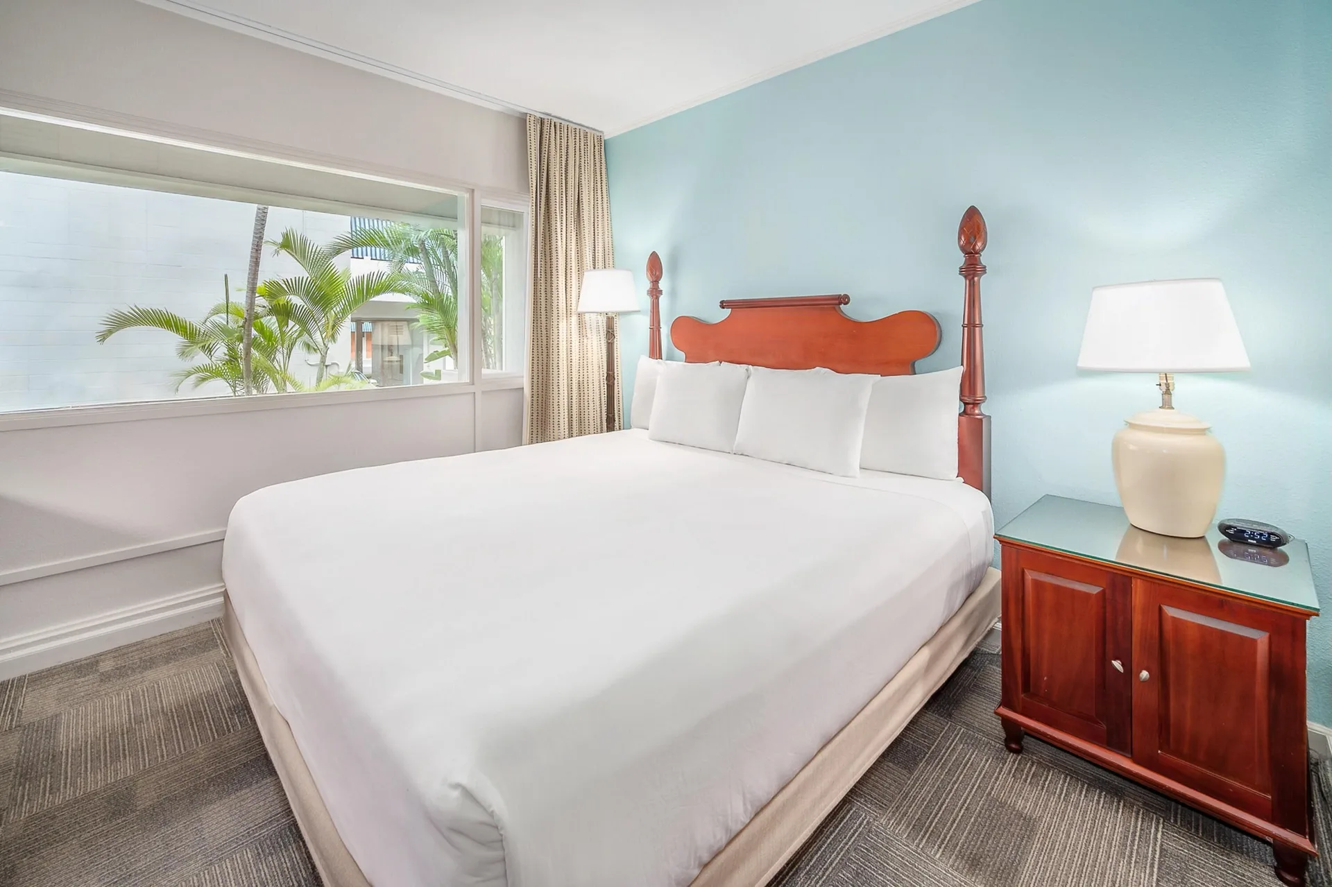 Standard Room One Bed at Maui Beach Hotel with white bed, blue wall, nightstand, lamps, and palm-facing window.