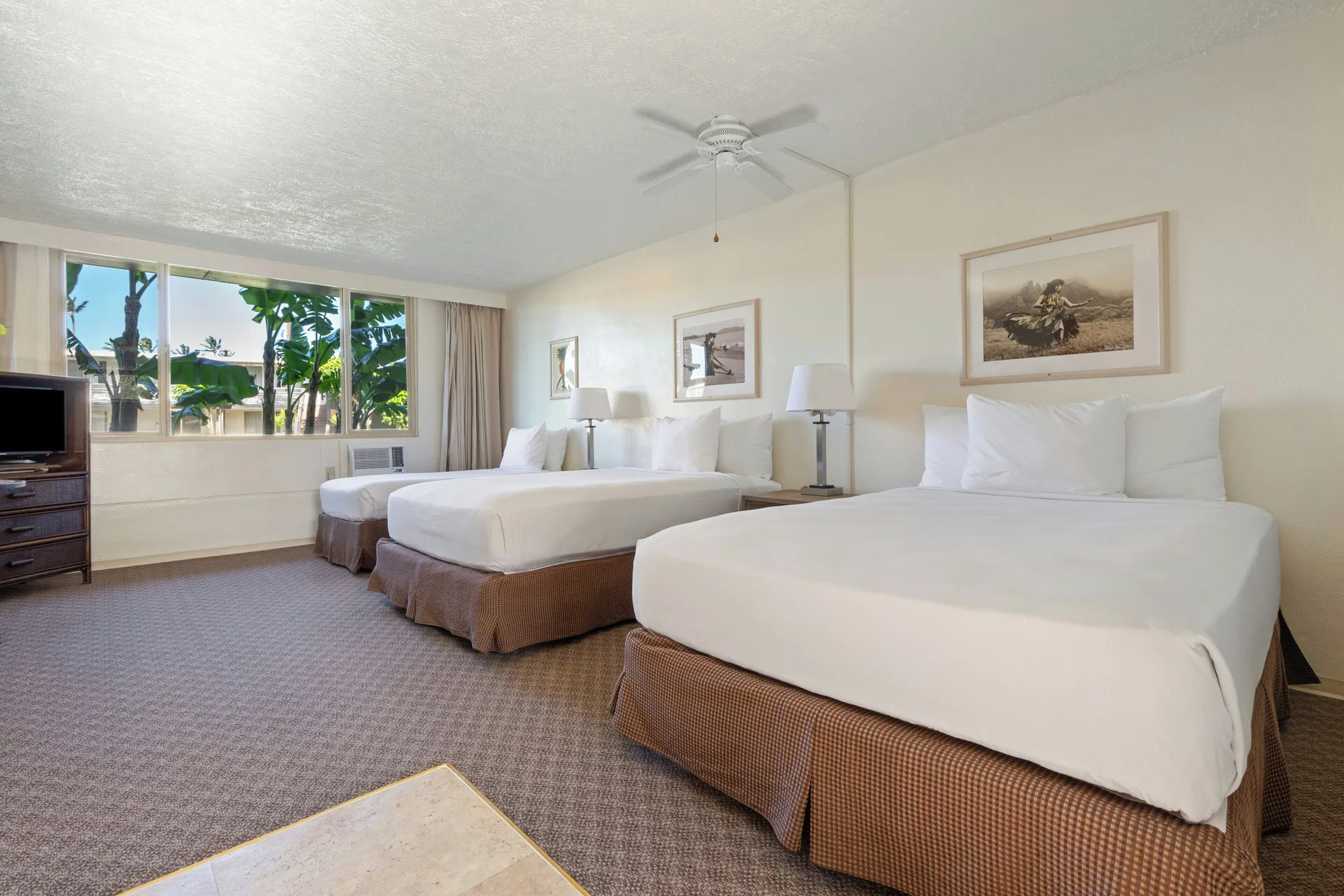 Poolside room with three beds, bedside lamps, TV, and tropical window view at Maui Seaside Hotel.