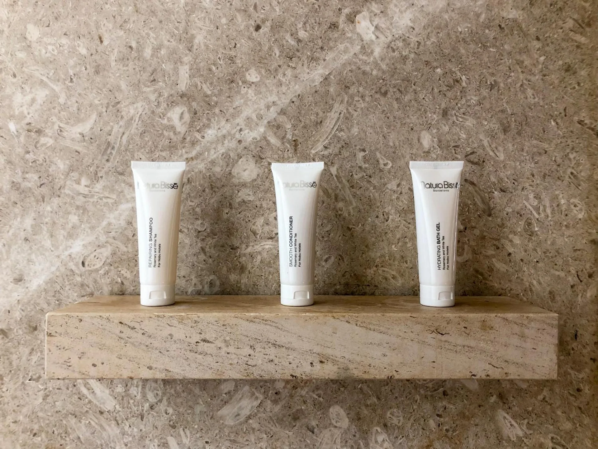 Three bath amenity tubes on a stone shelf in a deluxe room at Nobu Hotel Los Cabos.