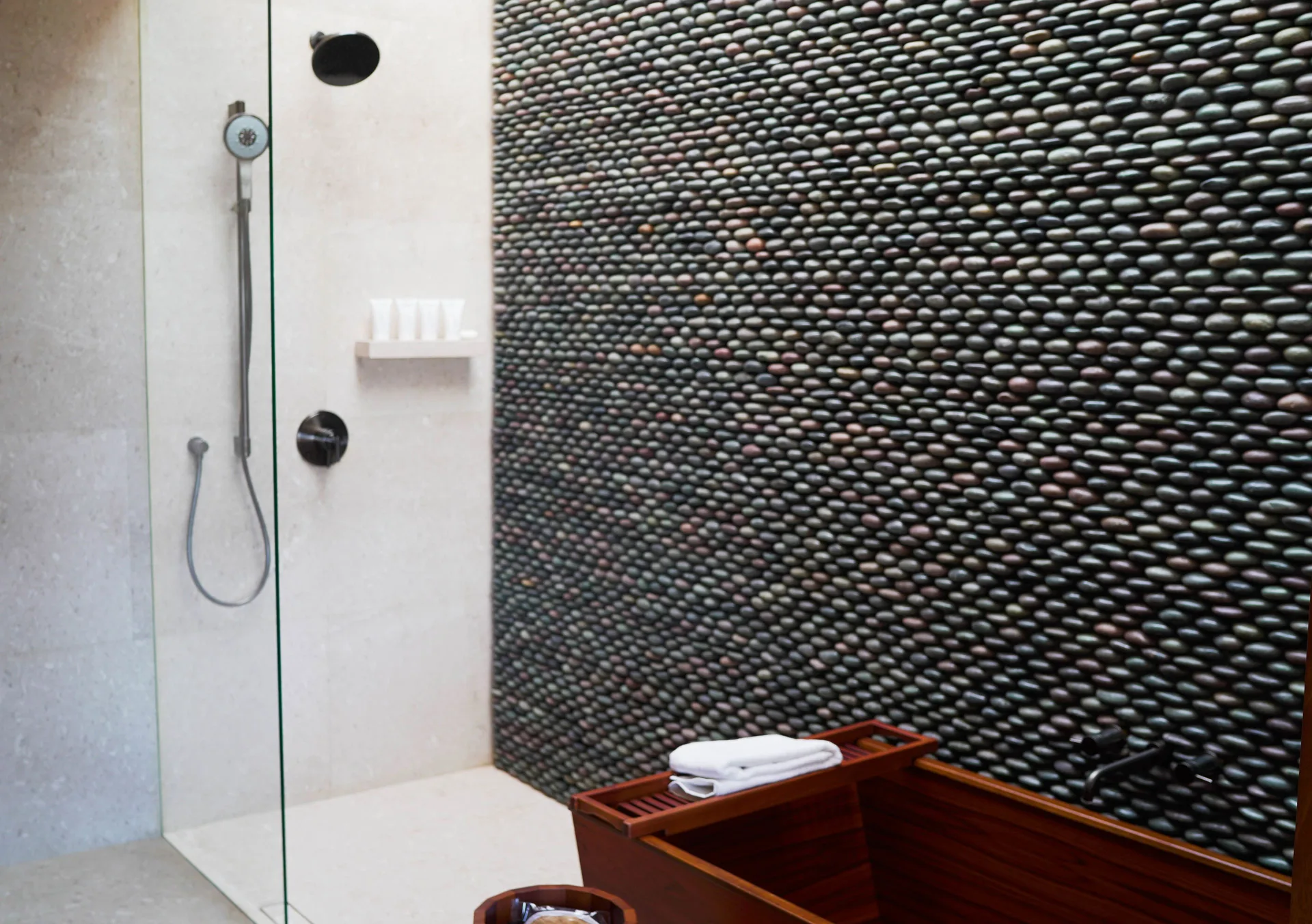 Junior Room shower with pebble wall, glass panel, and tub edge at Nobu Hotel Los Cabos.