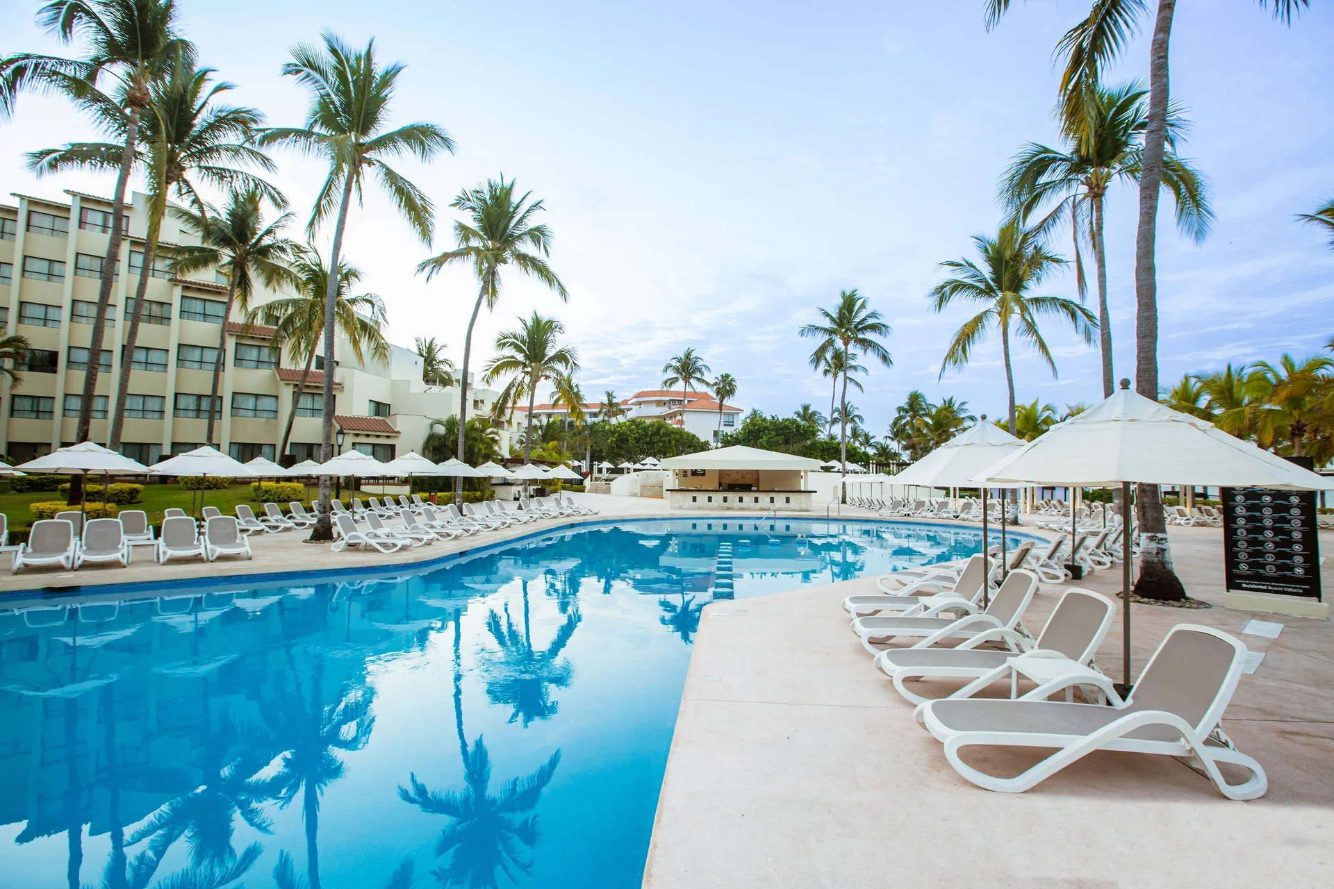 Pool deck with white loungers and umbrellas beneath tall palms at Occidental Nuevo Vallarta.