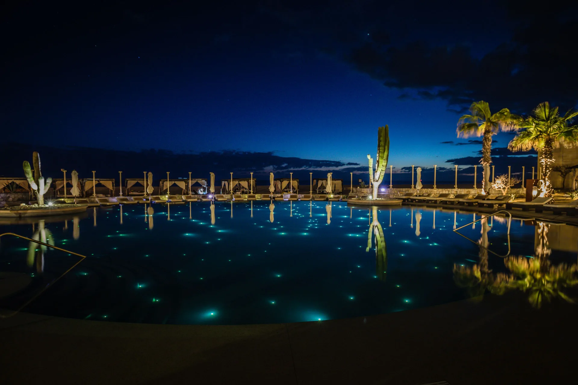 Illuminated pool water reflects palms and cabanas at Pueblo Bonito Pacifica Golf & Spa Resort at night.