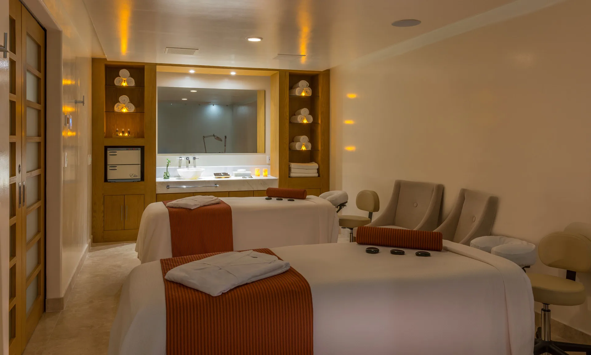 Spa treatment room with two massage tables, lounge chairs, shelves, and vanity at Pueblo Bonito Pacifica Golf & Spa Resort.