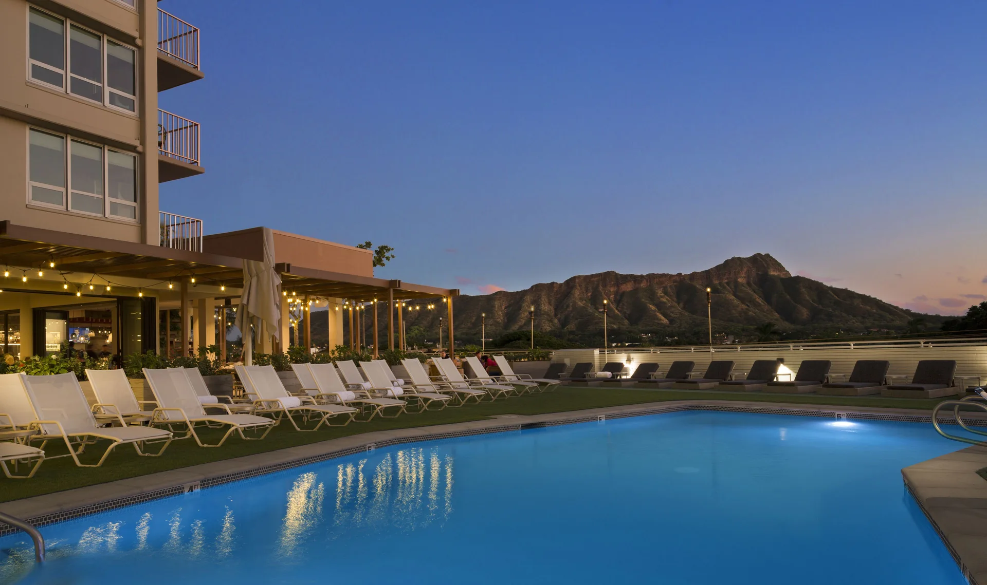 Evening pool deck with lit loungers and Diamond Head at Queen Kapiolani Hotel.
