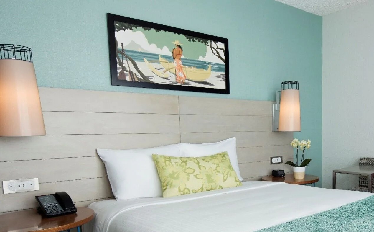 A king bed with bedside lamps and Hawaiian-style artwork at Queen Kapiolani Hotel.