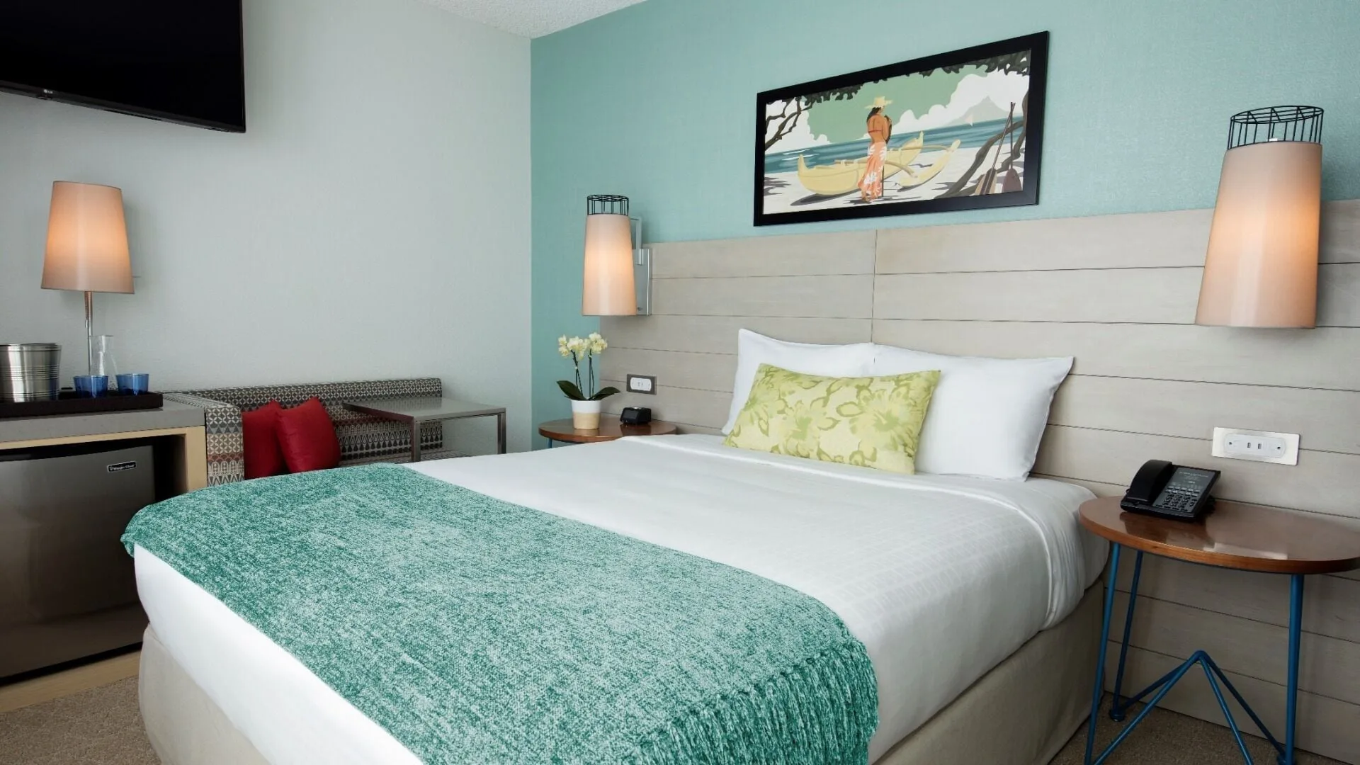 A king bed with turquoise throw and wall art fills a guest room at Queen Kapiolani Hotel.