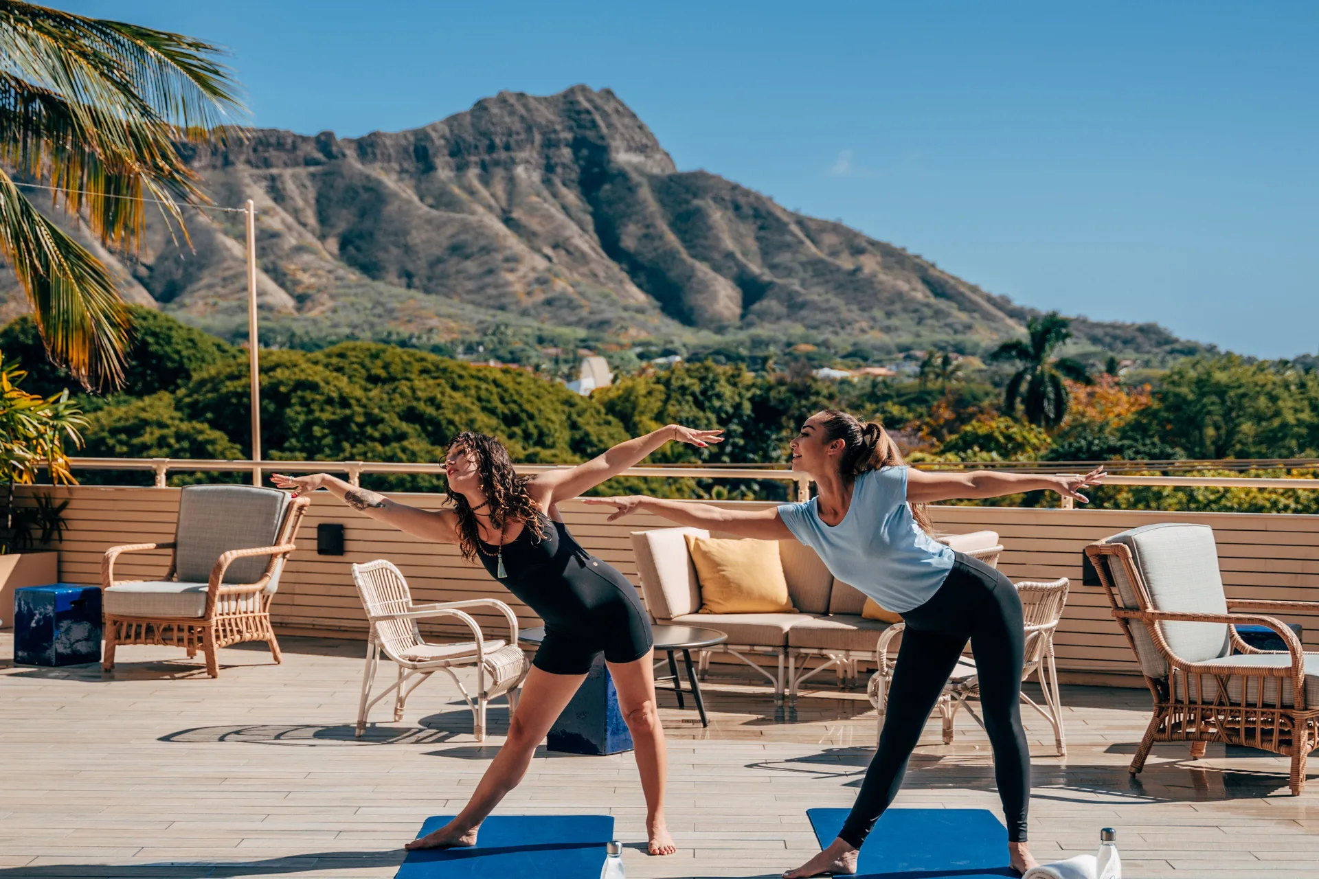 Two people practice yoga on an outdoor deck with Diamond Head beyond Queen Kapiolani Hotel.