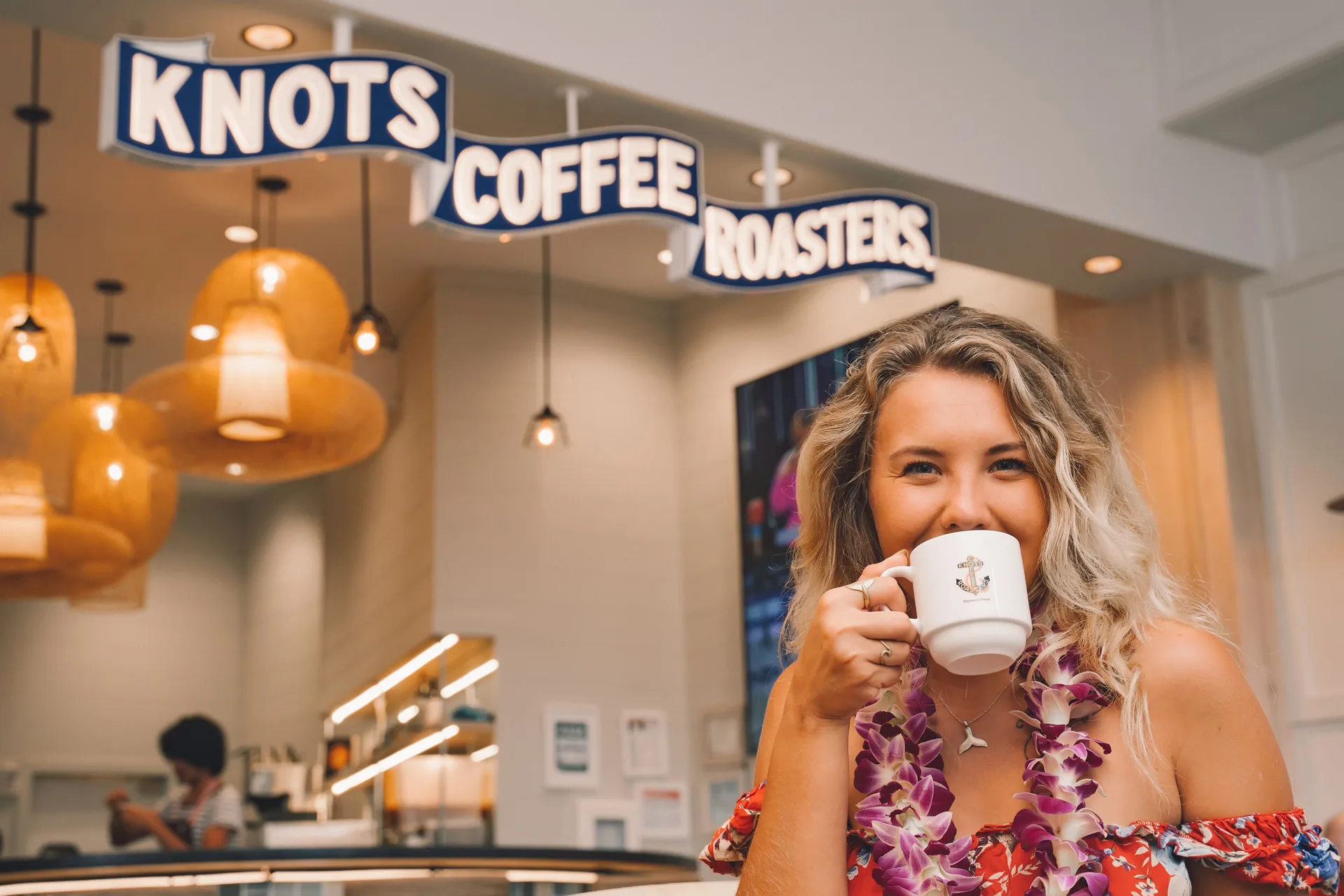 Guest holding a coffee mug beneath the Knots Coffee Roasters sign at Queen Kapiolani Hotel.