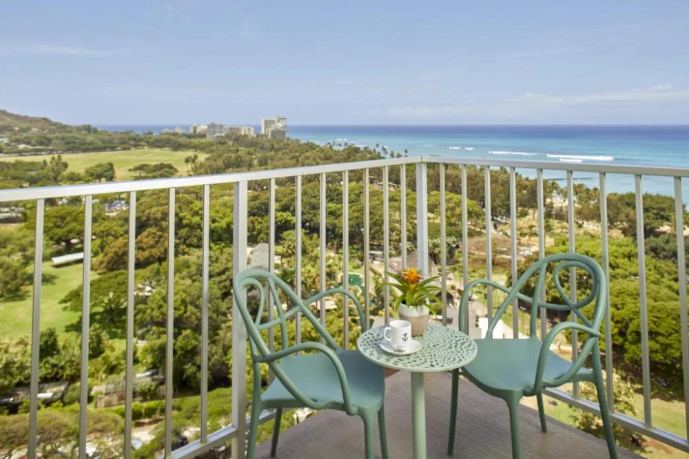Junior Suite Ocean View balcony seating overlooking ocean and park at Queen Kapiolani Hotel.