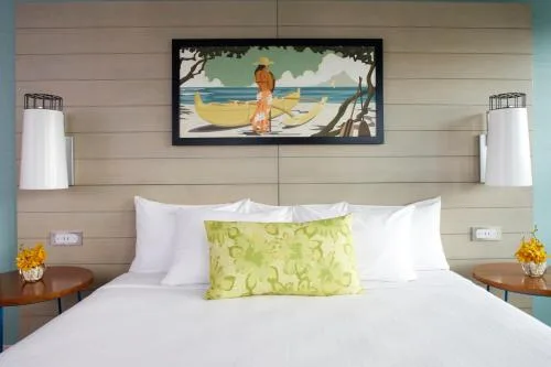 Studio king bed with coastal art, white pillows, and bedside lamps at Queen Kapiolani Hotel.