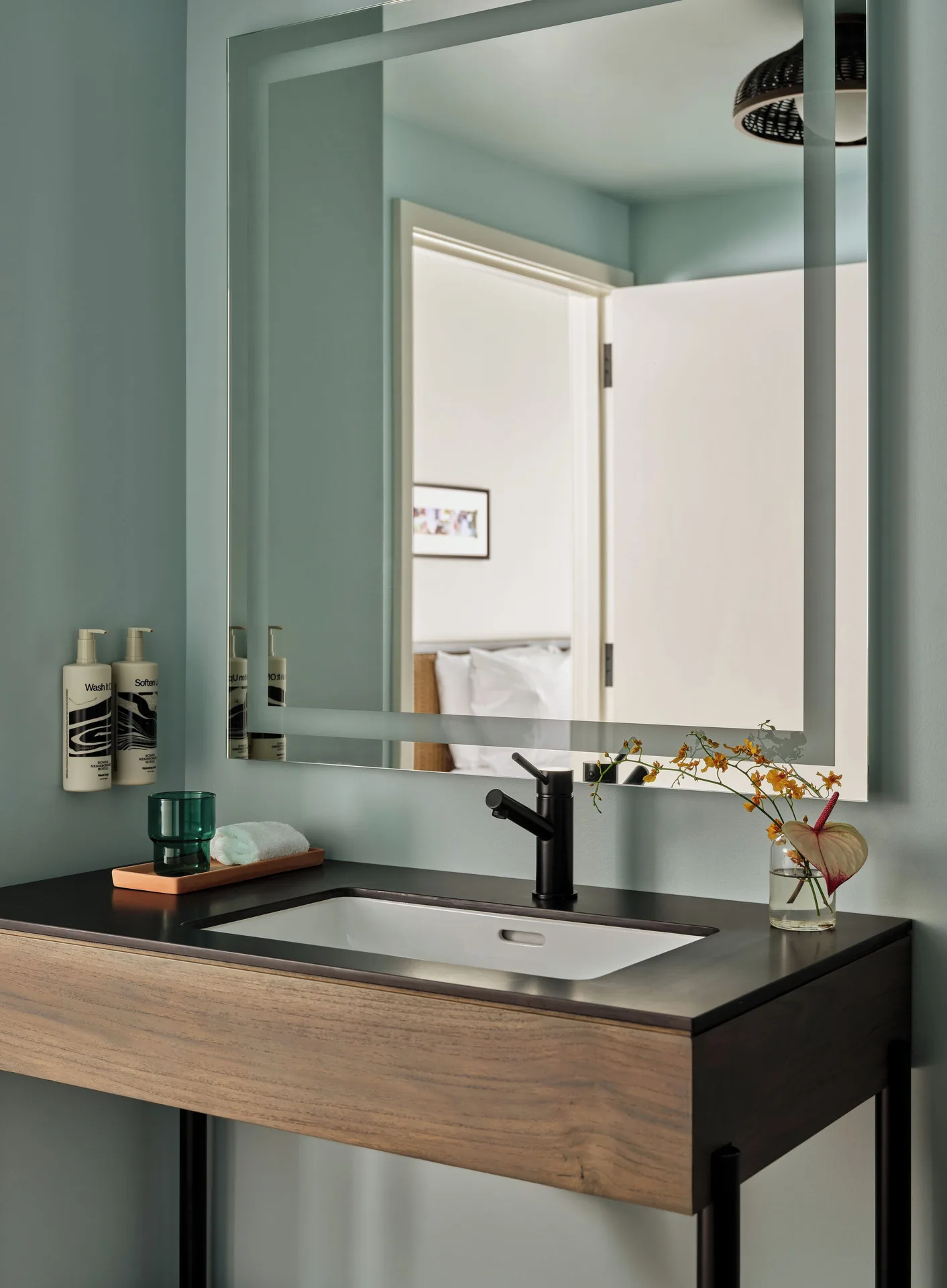 Standard room bathroom vanity at Romer House Waikiki with black faucet, large mirror, and flower vase.